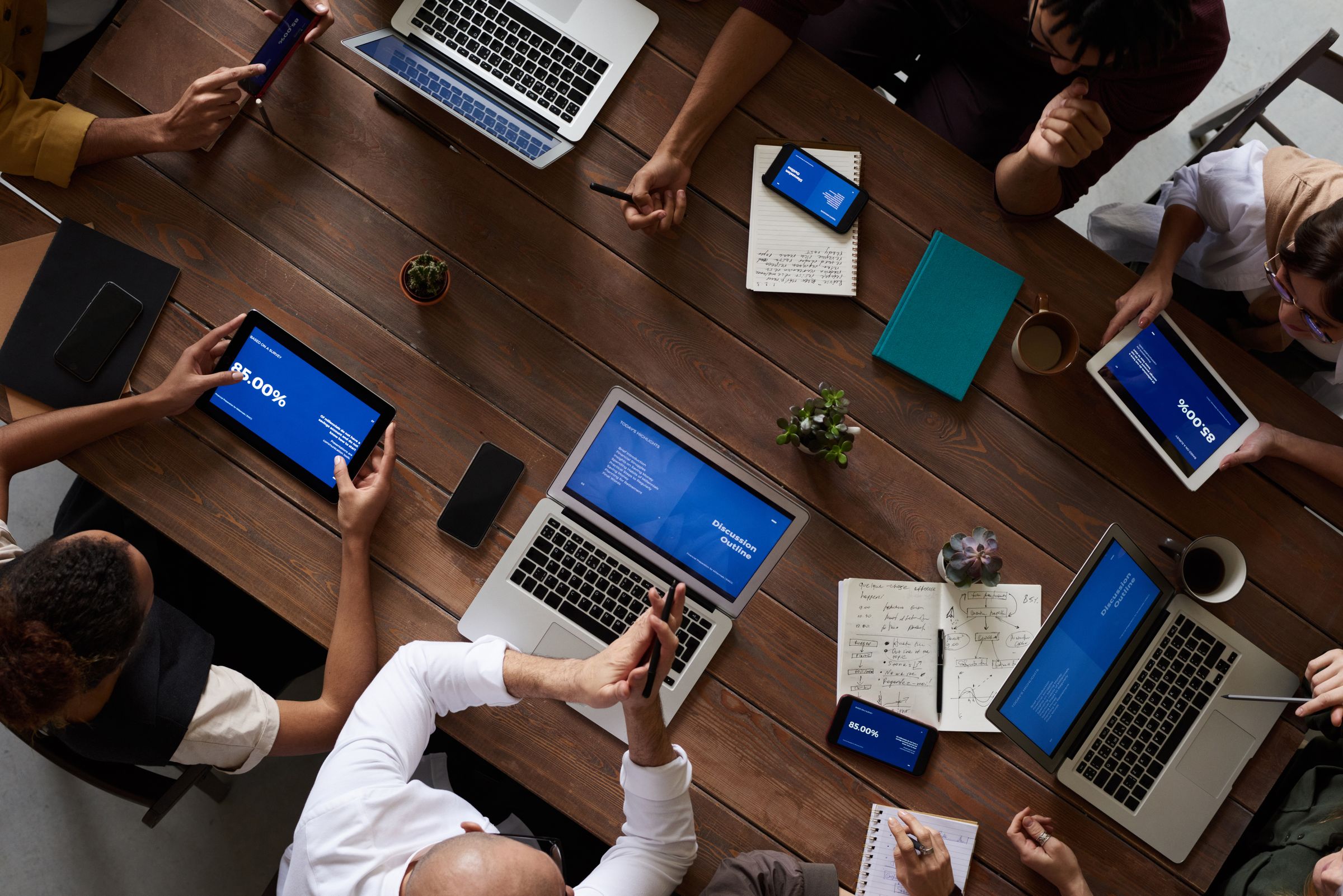 An overhead view of colleagues around a dark wooden table with laptops, tablets and notebooks.