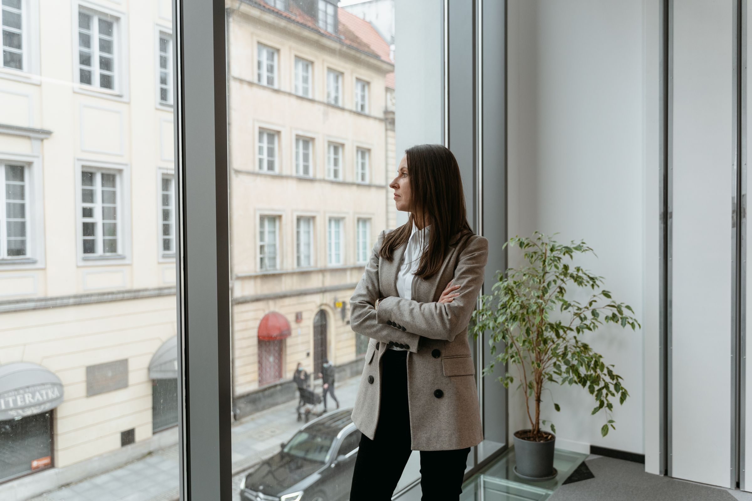 A woman in a blazer standing with folded arms at a tall office window looking out on a street of old buildings.
