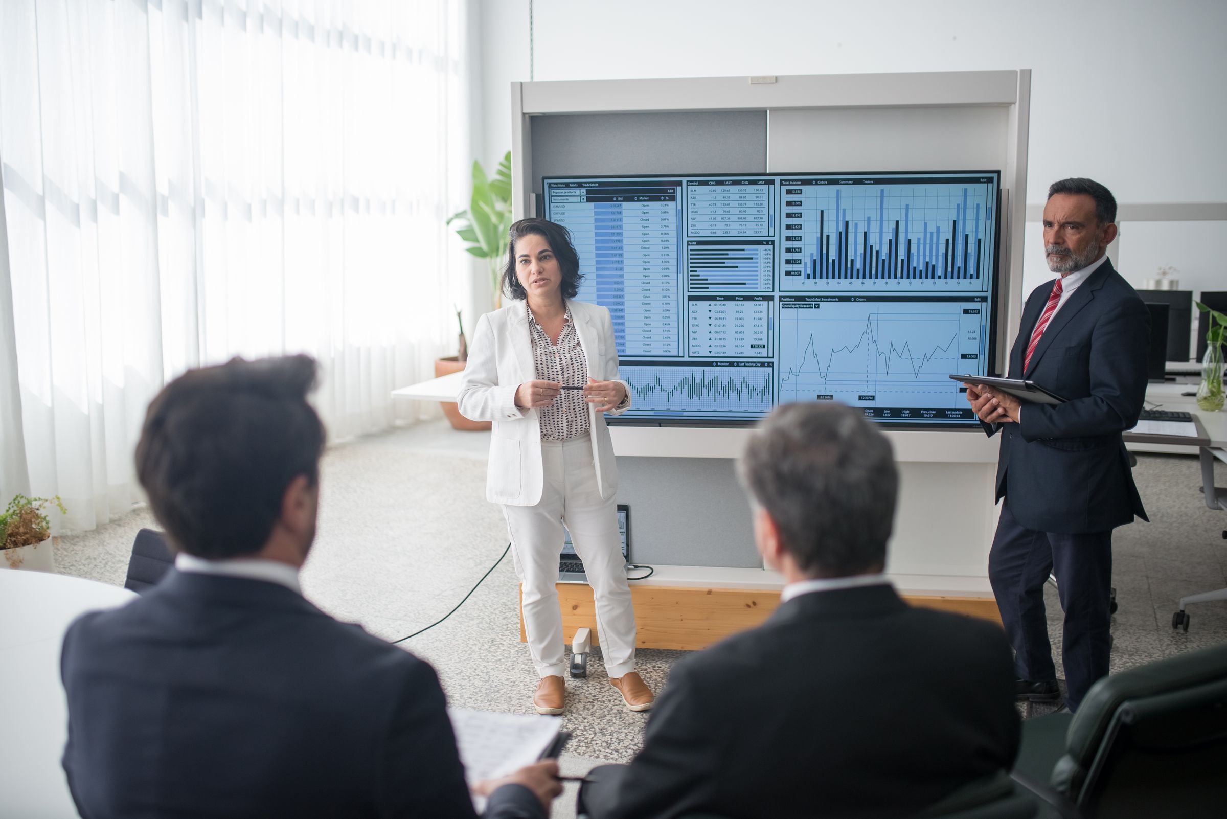 A presenter standing beside a large screen of charts while colleagues watch from behind.