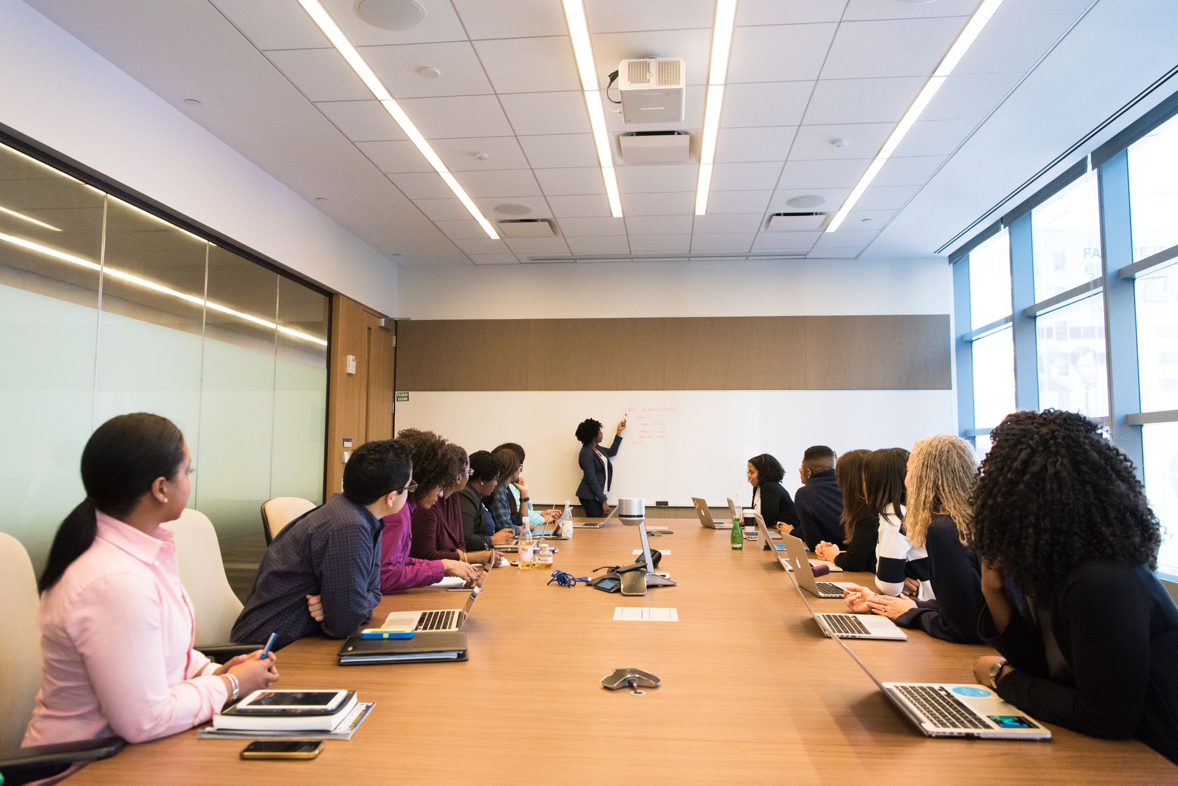 A group seated along a long wooden meeting table while one person stands and points to a screen at the far end.
