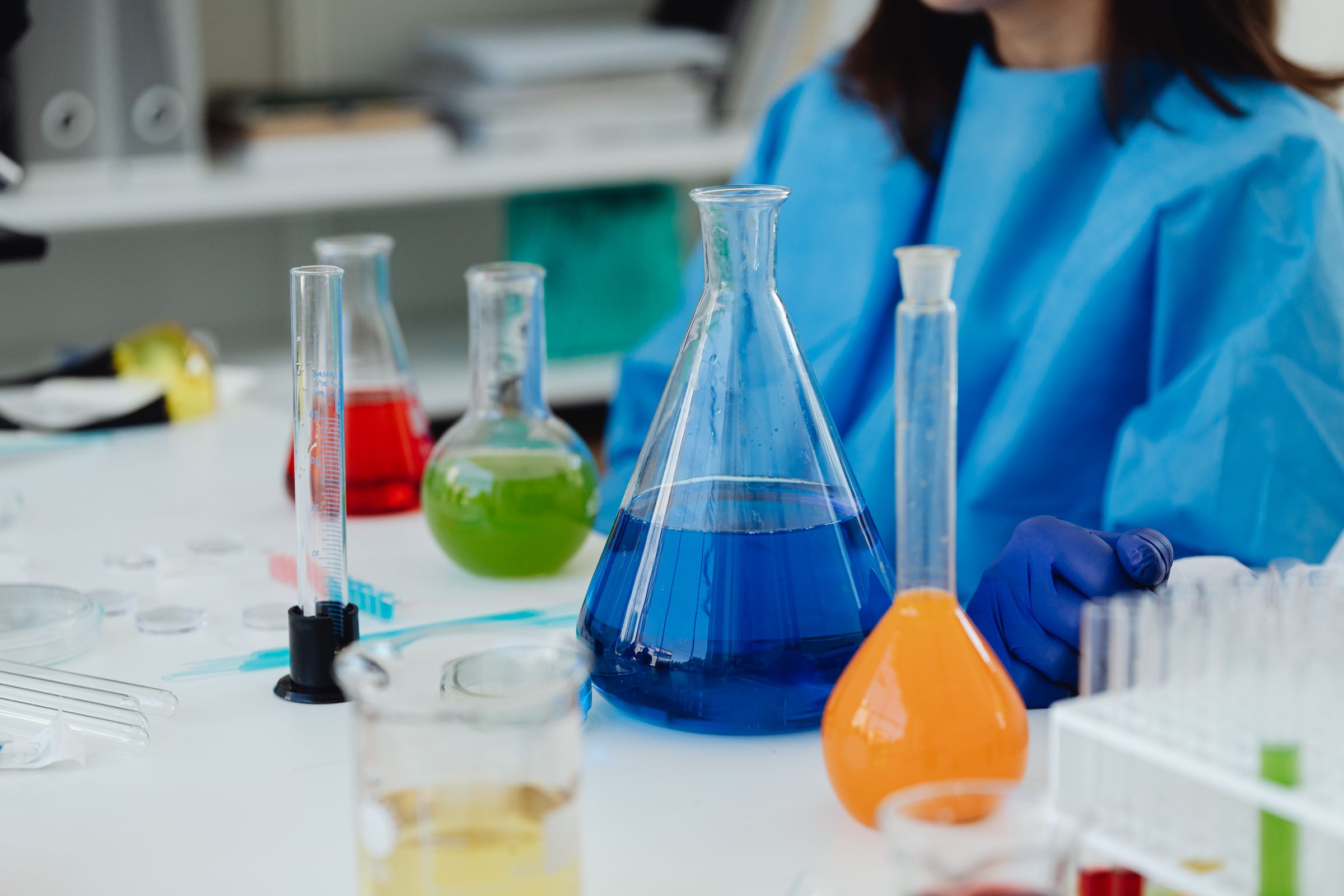 A laboratory bench with flasks of blue, orange and green liquid, and a researcher in blue protective clothing working behind them.
