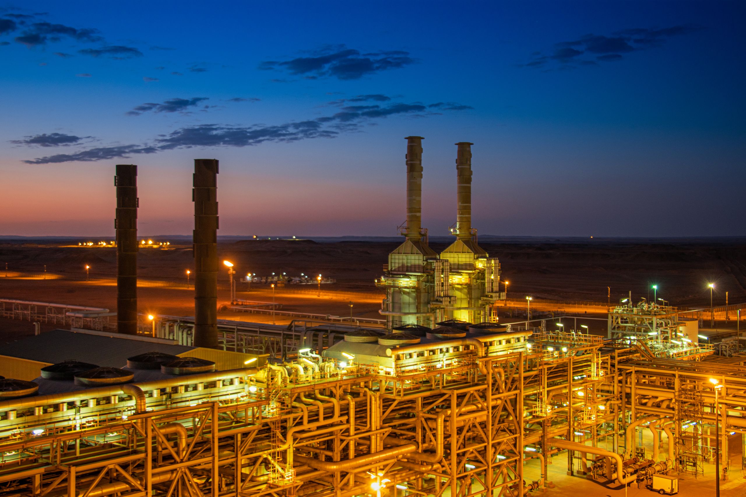 An industrial chemical plant at dusk, with lit pipework and stacks glowing orange against a deep blue sky.