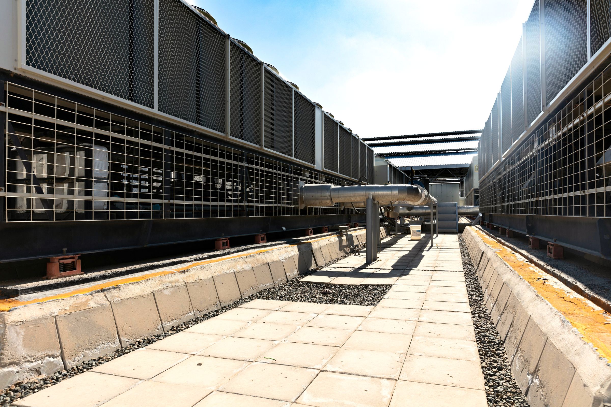 A paved walkway between two long banks of industrial cooling units on a plant roof under a bright sky.