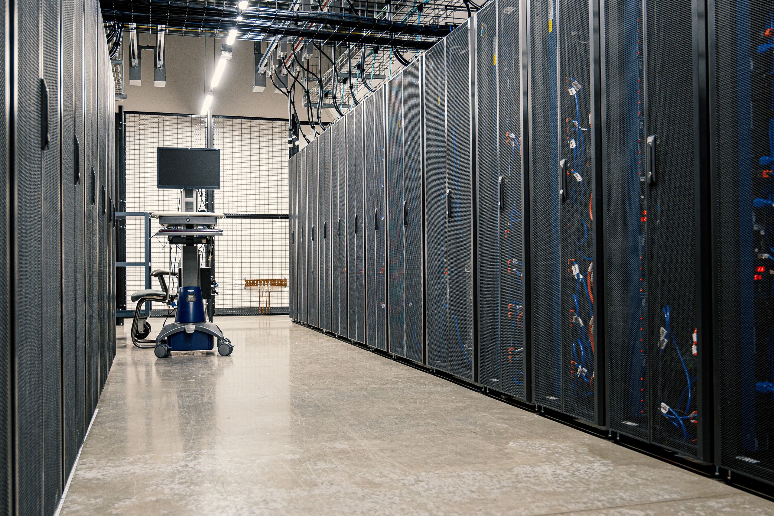 A long aisle between tall dark server cabinets in a data hall, with cable trays and strip lights overhead.