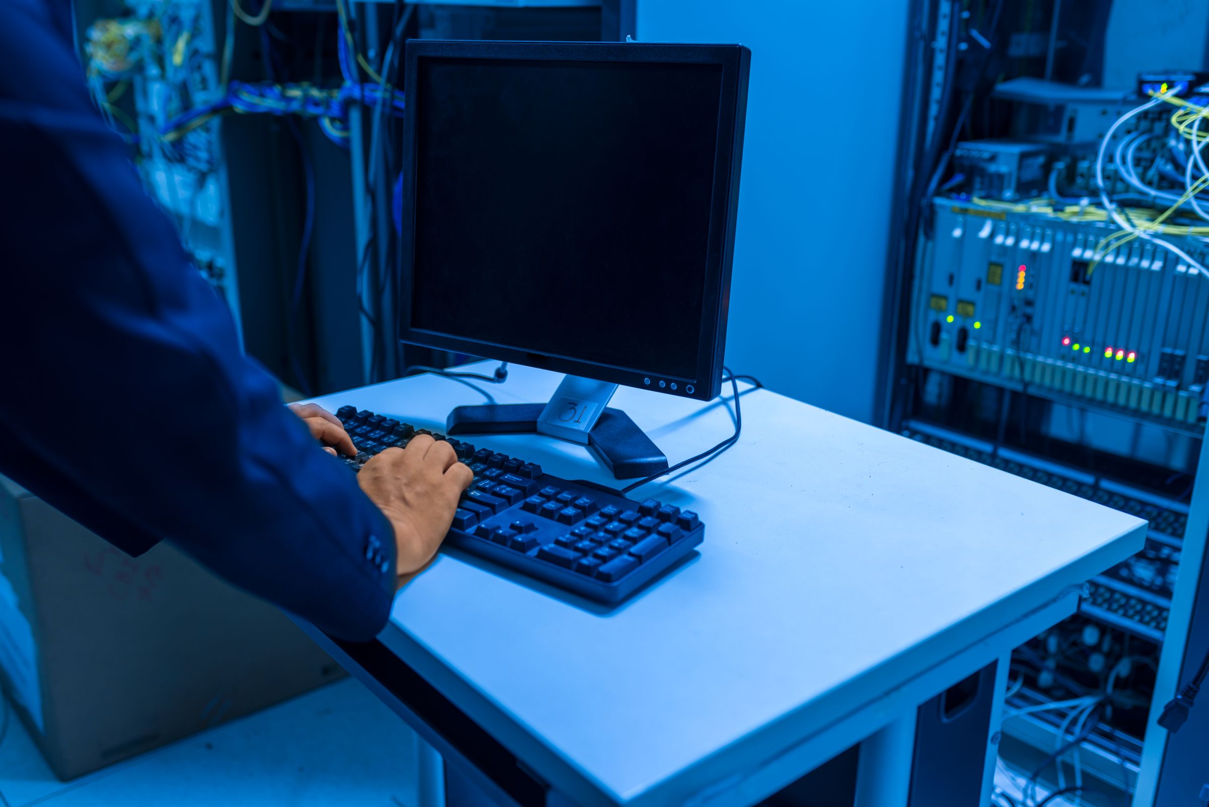 A technician's hands typing at a monitor and keyboard beside a lit, cabled equipment rack.