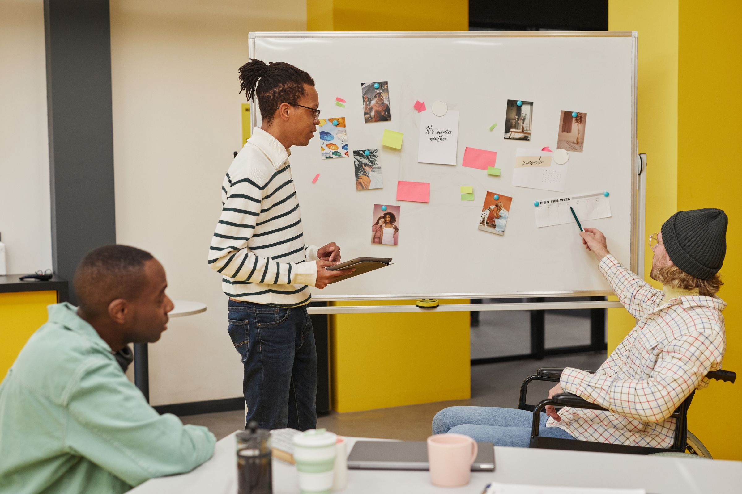 Three colleagues at a table with a whiteboard of pinned photographs and sticky notes behind them.