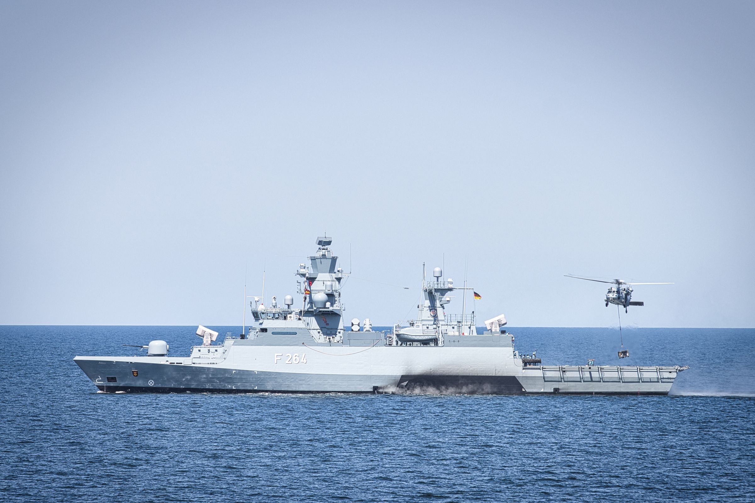 A naval frigate at sea with a helicopter hovering alongside