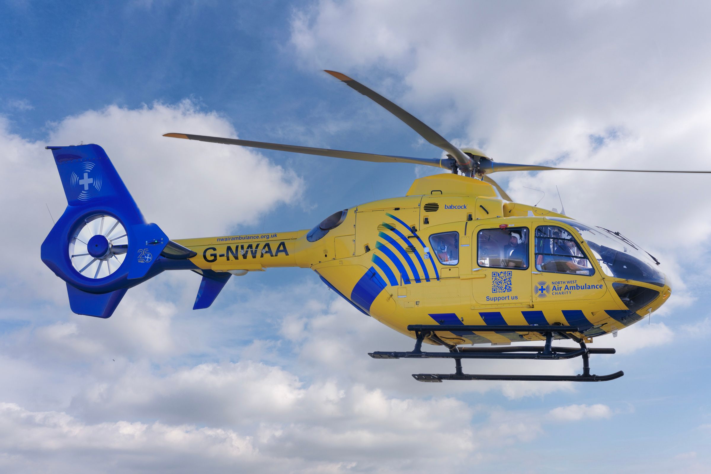 A yellow and blue air ambulance helicopter in flight against a sky with broken cloud.