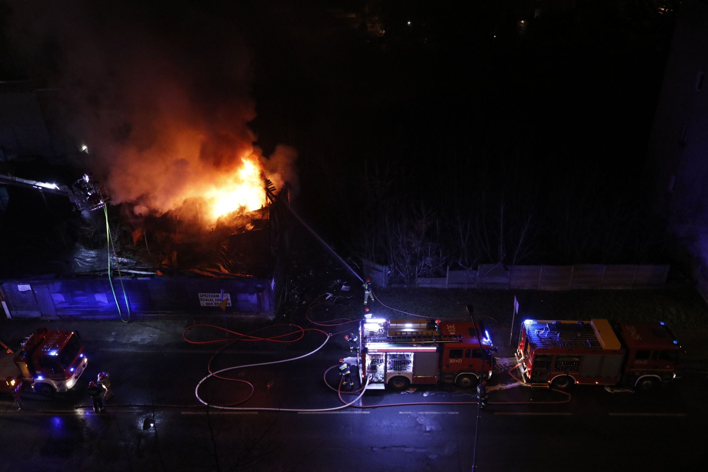 Fire engines parked on a street at night with hoses laid out, and a building burning behind them.