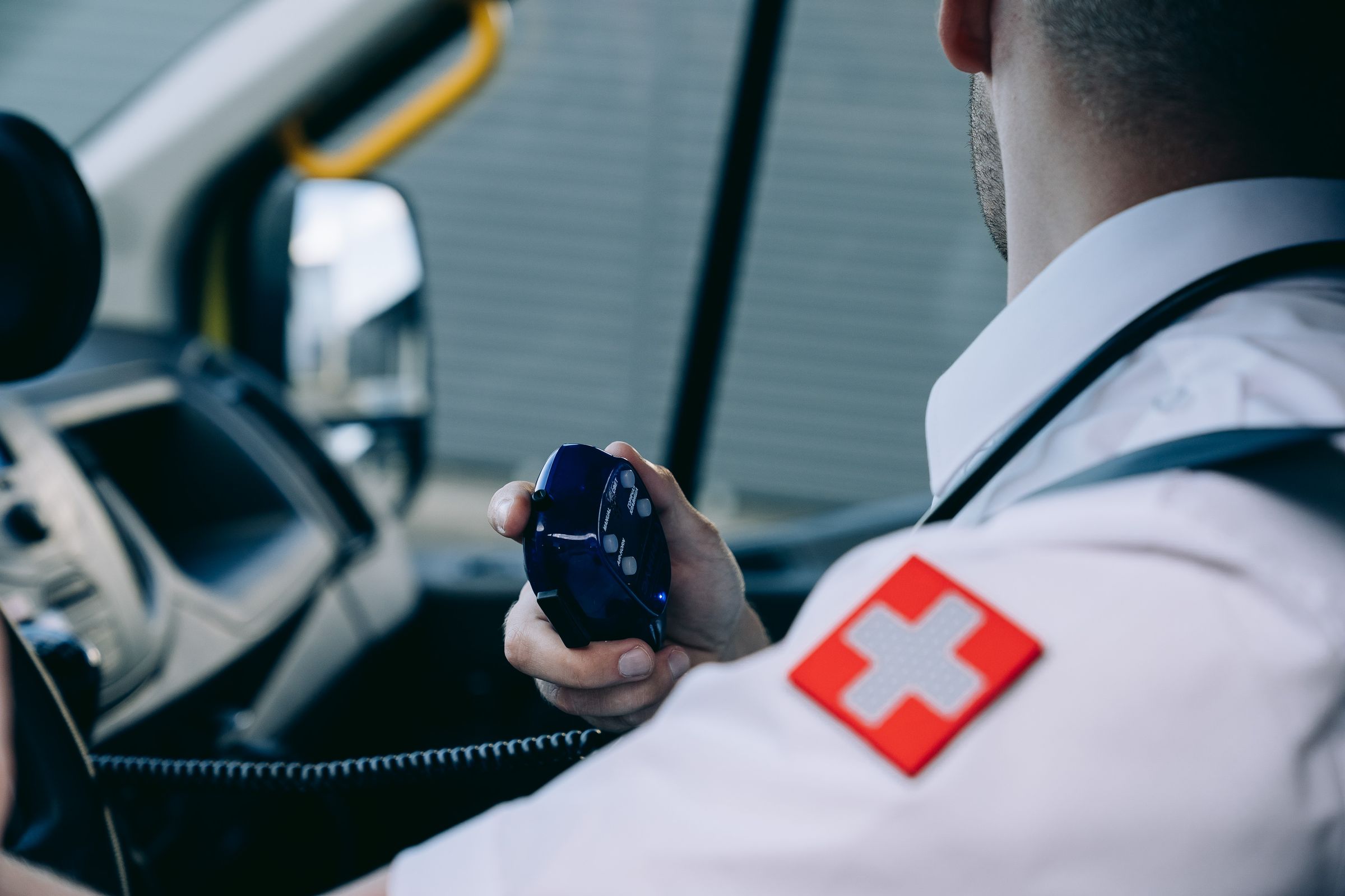 A paramedic in a white uniform holding a handheld radio inside an ambulance cab.