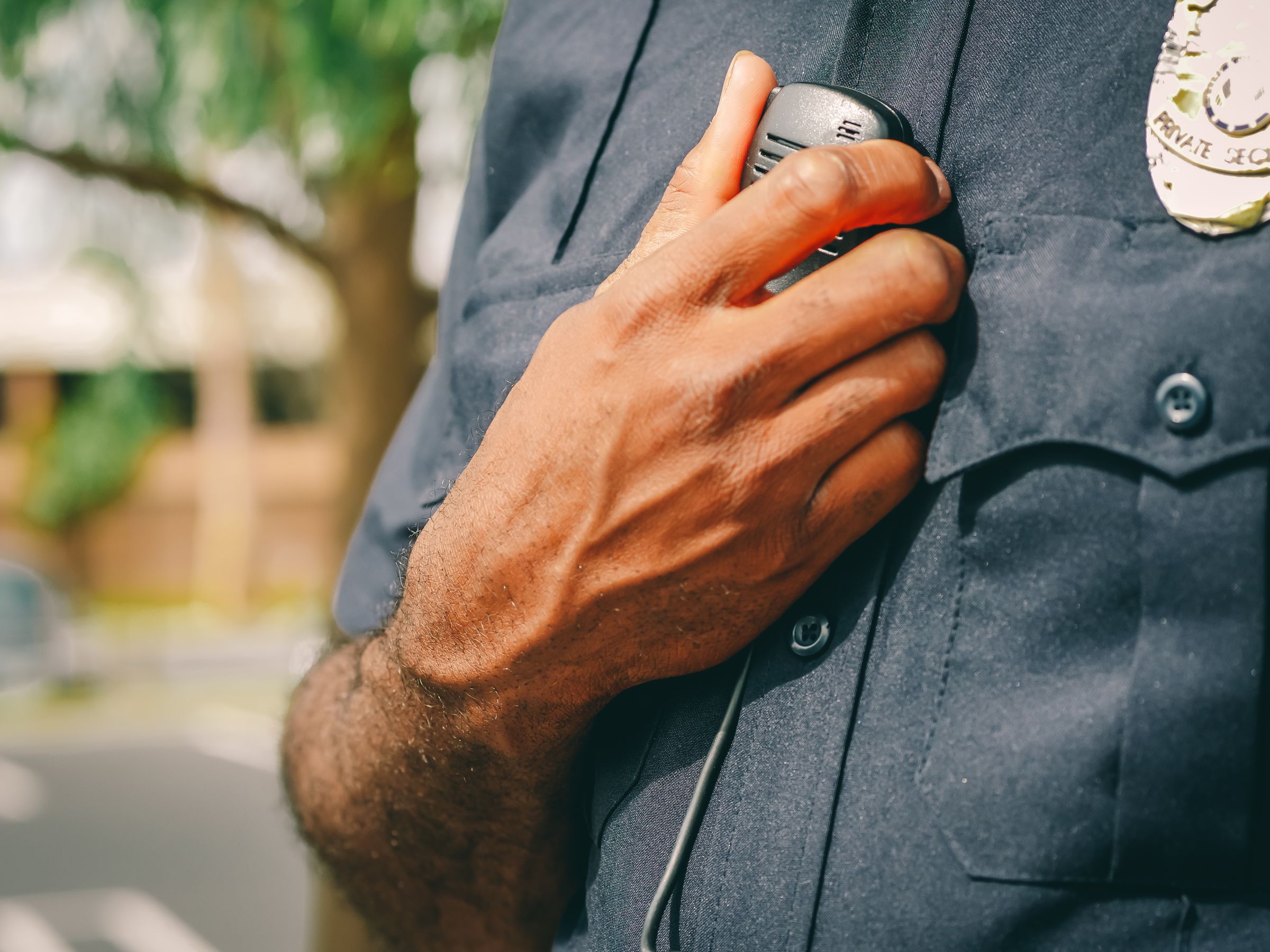 A close-up of a police officer's hand holding the radio clipped to the chest of a dark uniform.