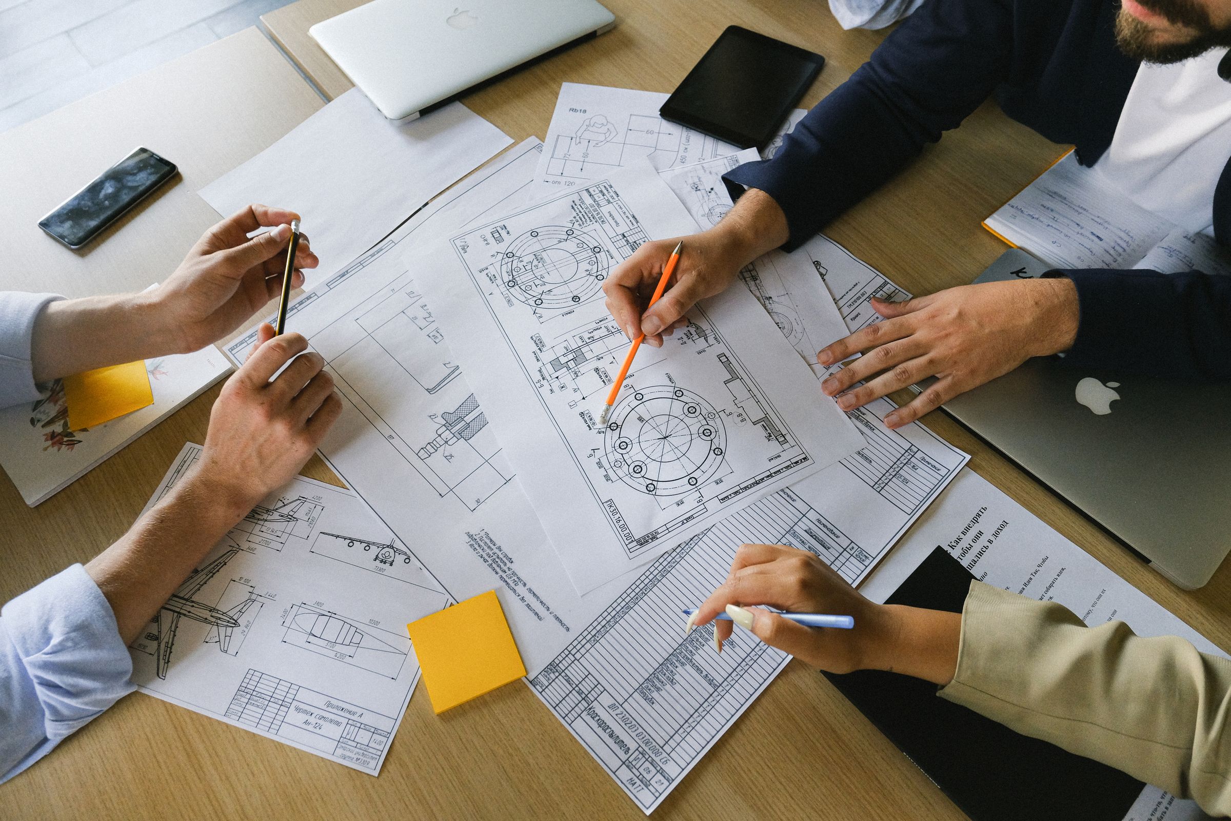 Hands holding pencils over engineering drawings and sticky notes on a wooden table, seen from above.