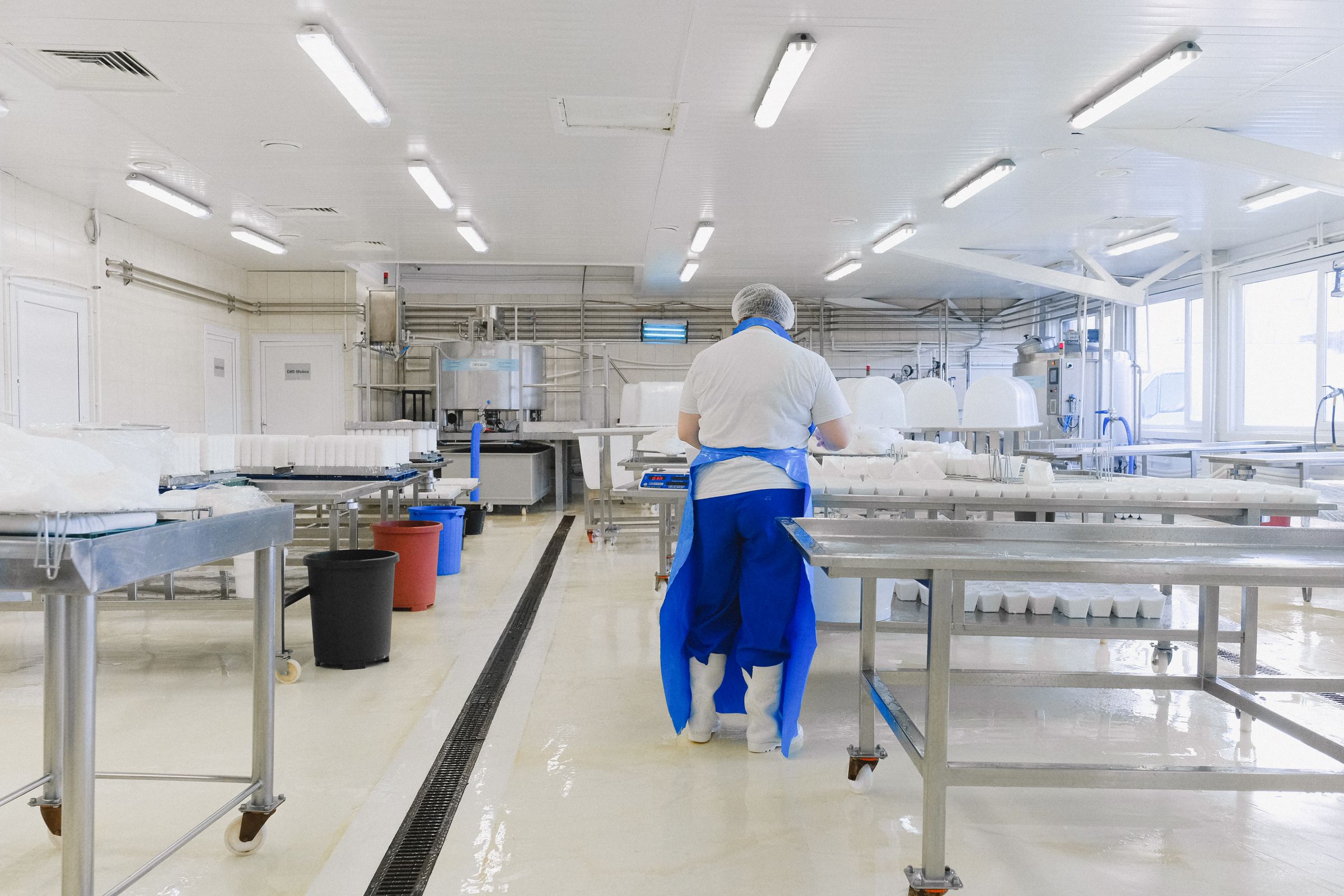 A worker in a blue apron and hairnet at a stainless steel table in a bright food preparation room, seen from behind.