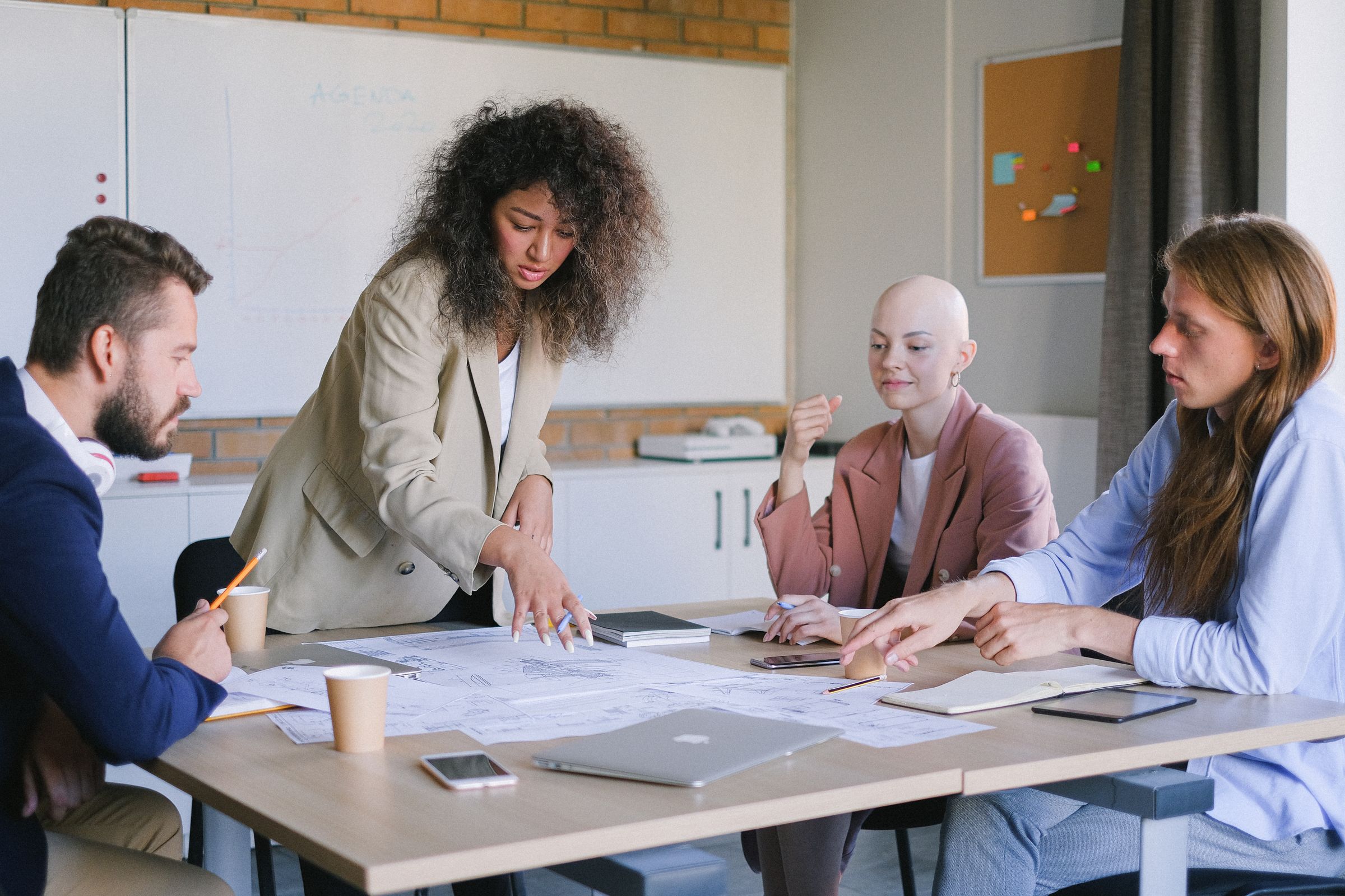 Four colleagues around a light wooden table, one standing and pointing at printed plans while the others look on.