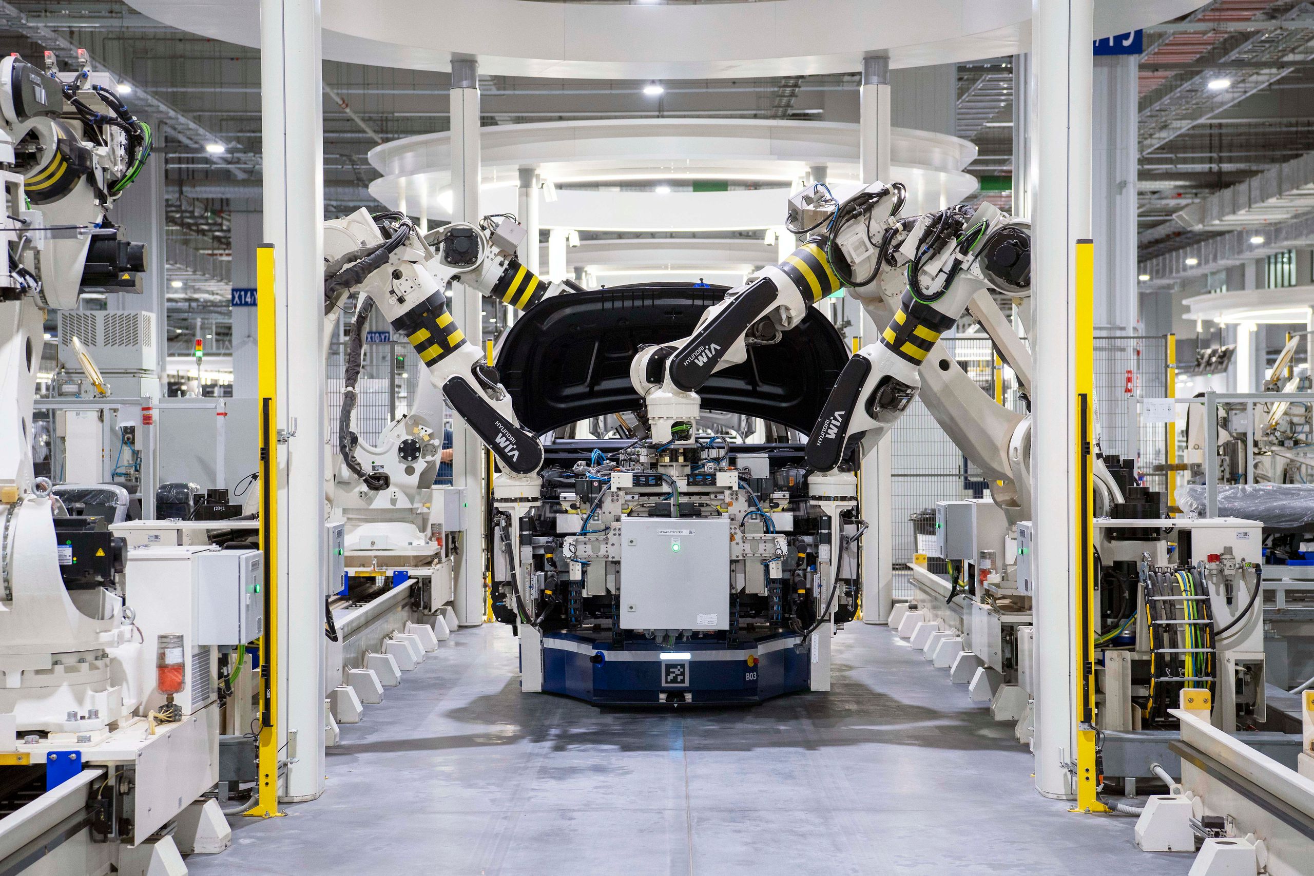 Robot arms assembling a vehicle body on an automated production line in a large, bright factory hall.