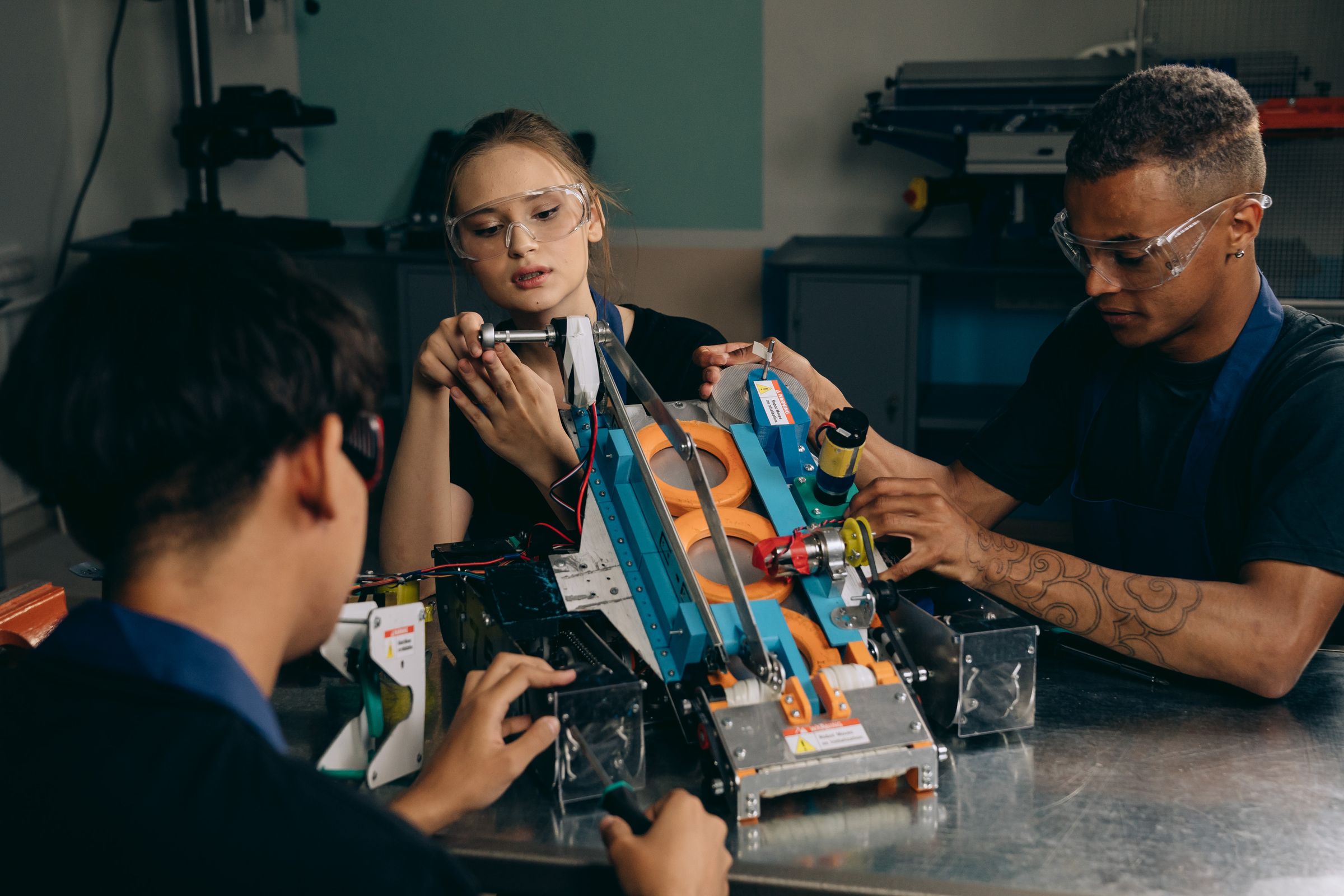 Three people in safety glasses working together on a small robotic vehicle at a workshop bench.