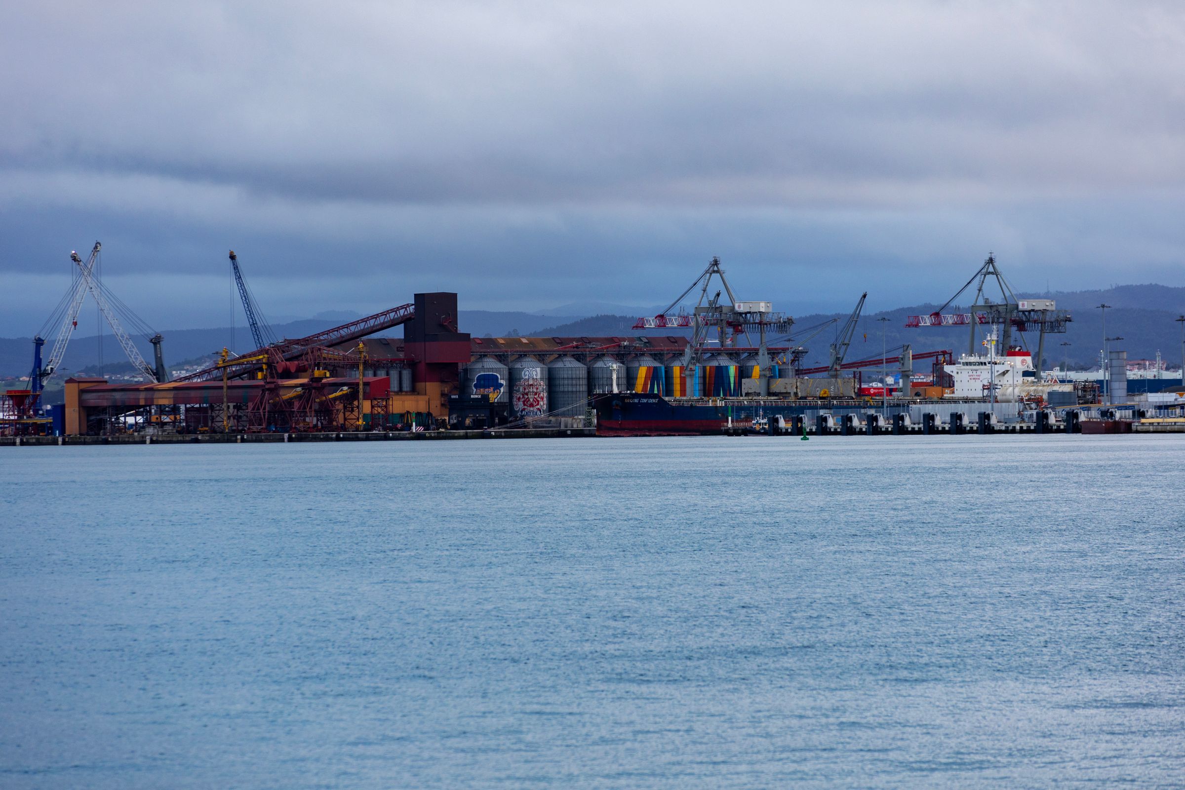 A harbour across grey-blue water, with quayside cranes and loading equipment under an overcast sky.