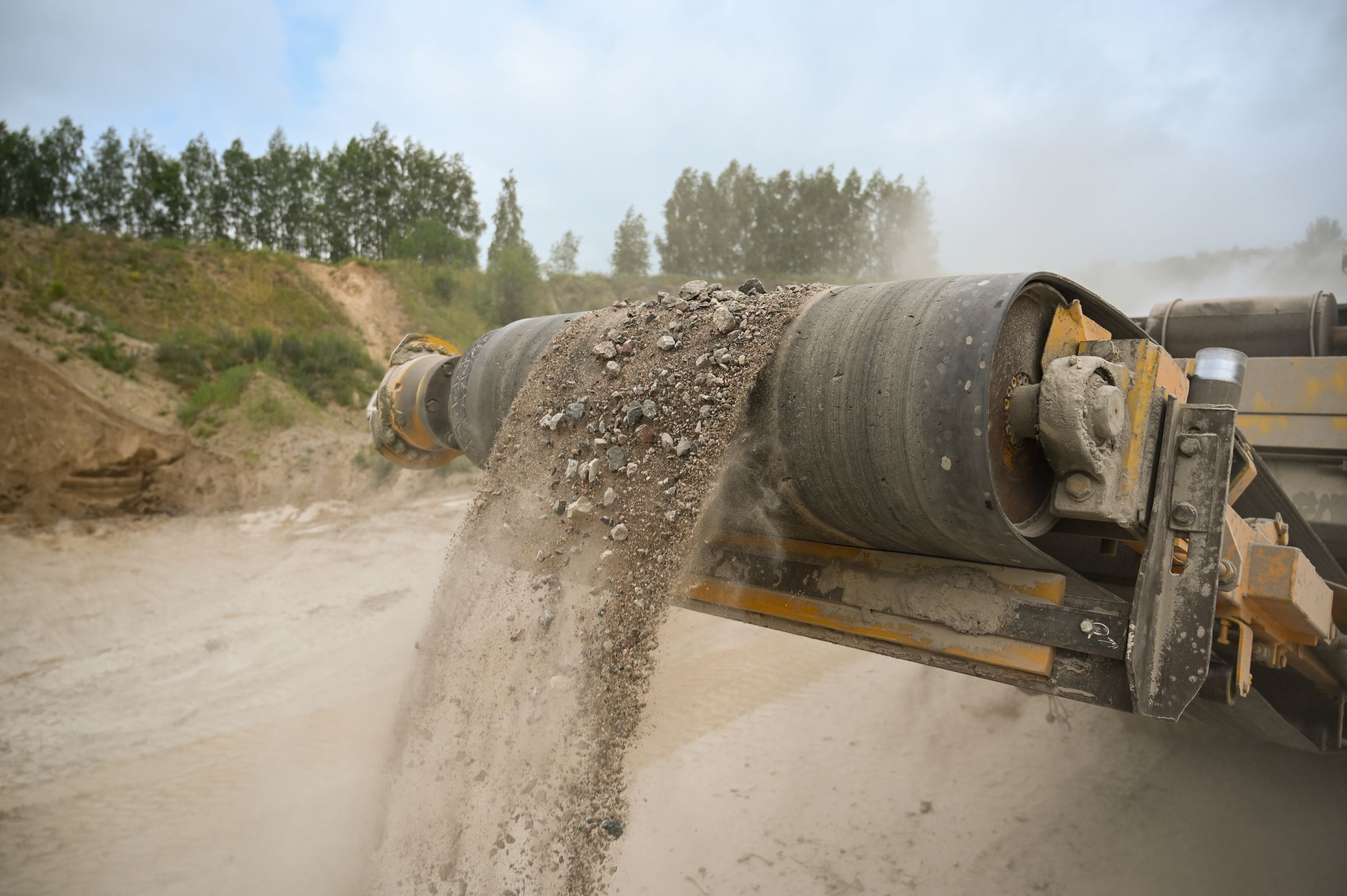 A conveyor belt carrying crushed rock out of a processing unit at a quarry, with dust in the air.