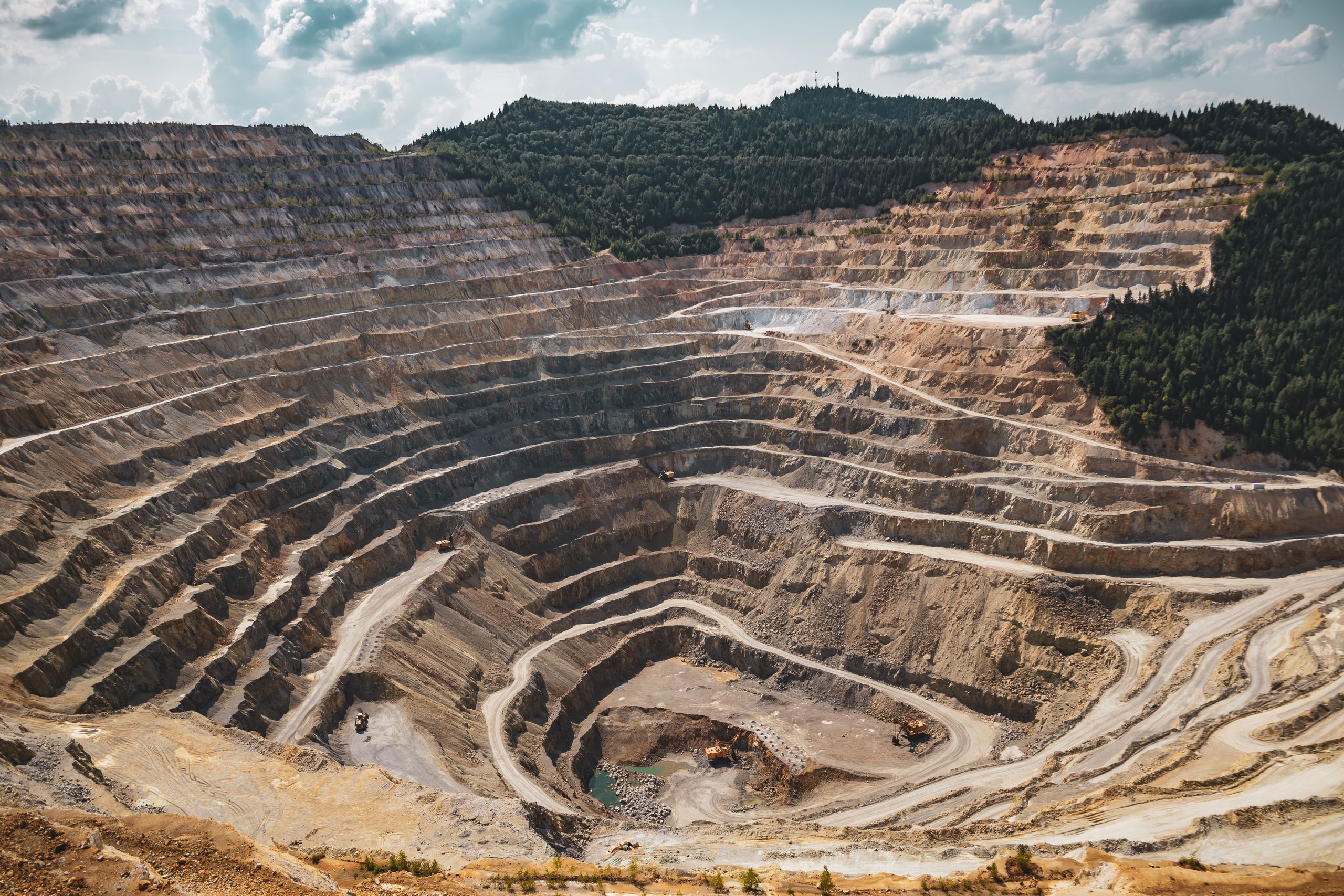 A terraced open-pit mine with winding haul roads cut into the bench walls beneath a partly cloudy sky.