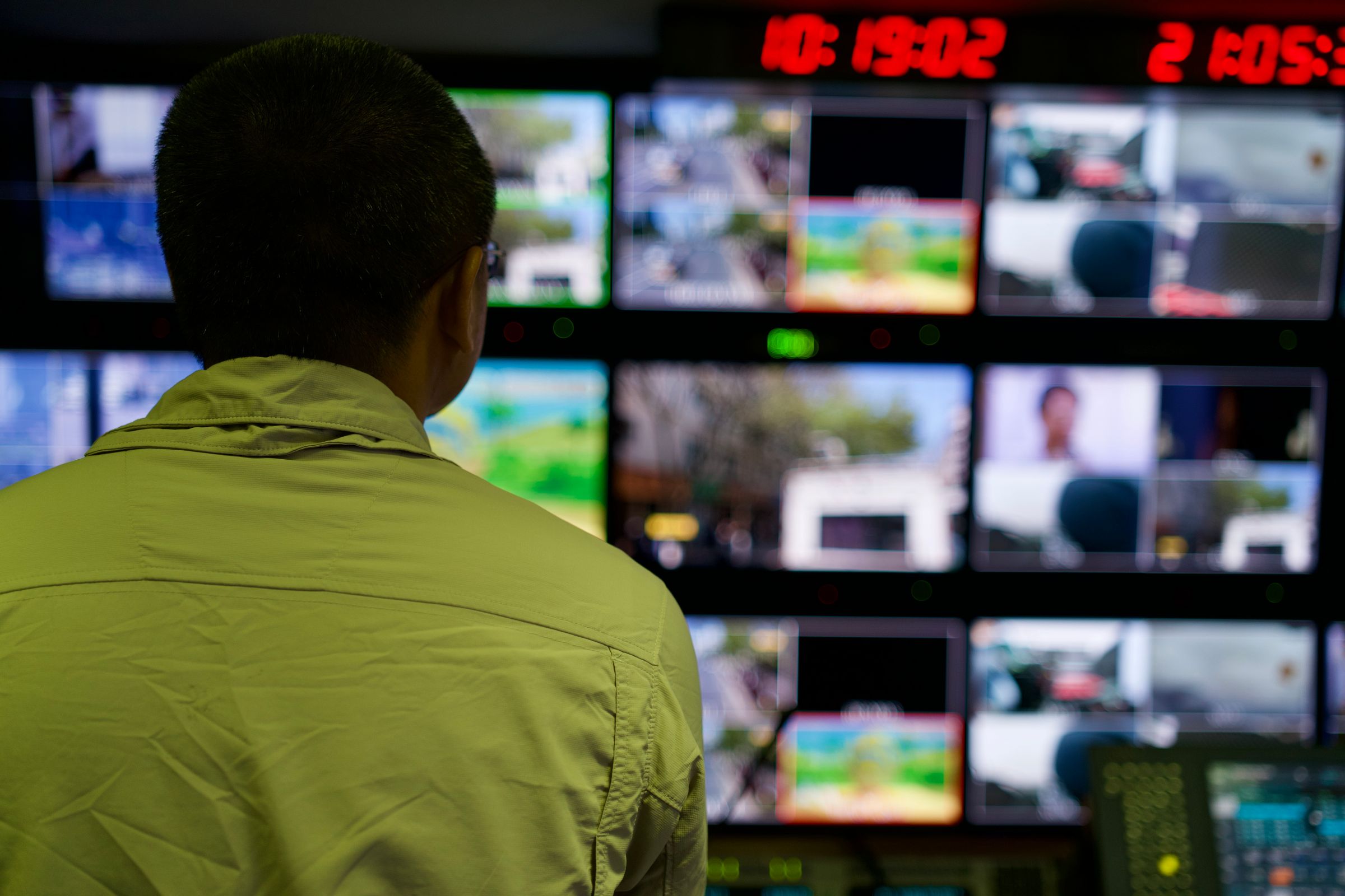 A man in a green jacket, seen from behind, watching a wall of monitors in an operations room.