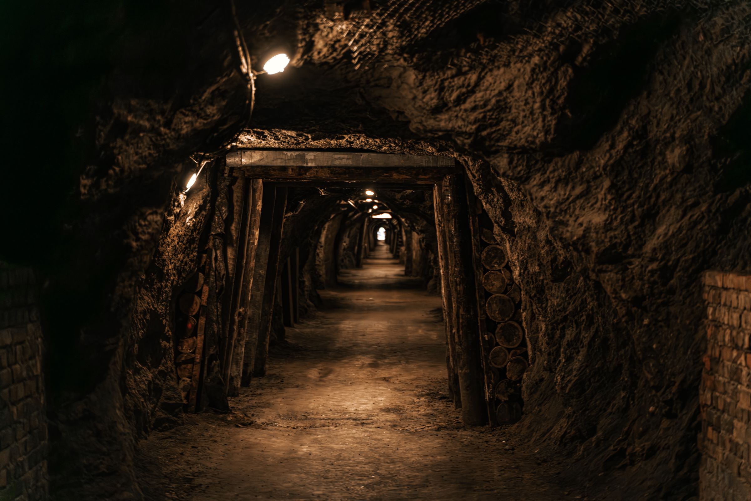 A timber-framed underground mine tunnel receding into darkness, lit by a single lamp.
