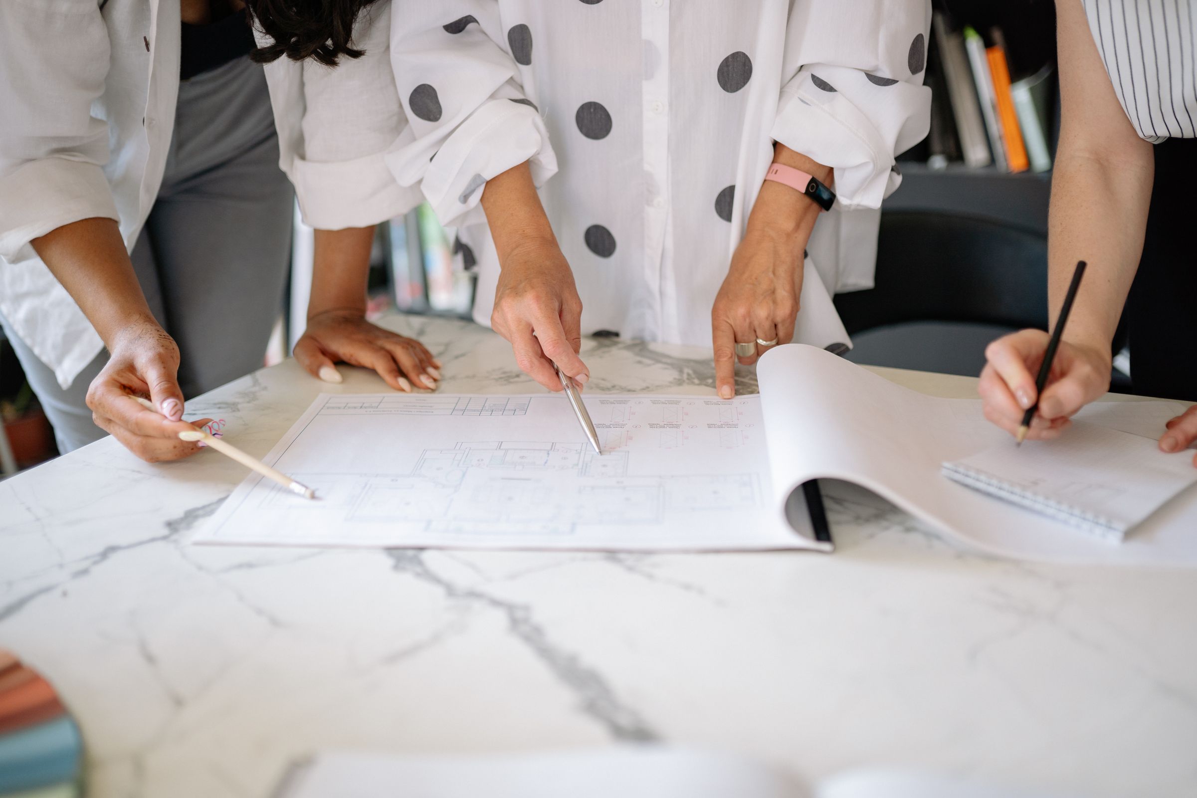 Hands of several colleagues resting on large technical drawings spread across a table, with pencils in hand.