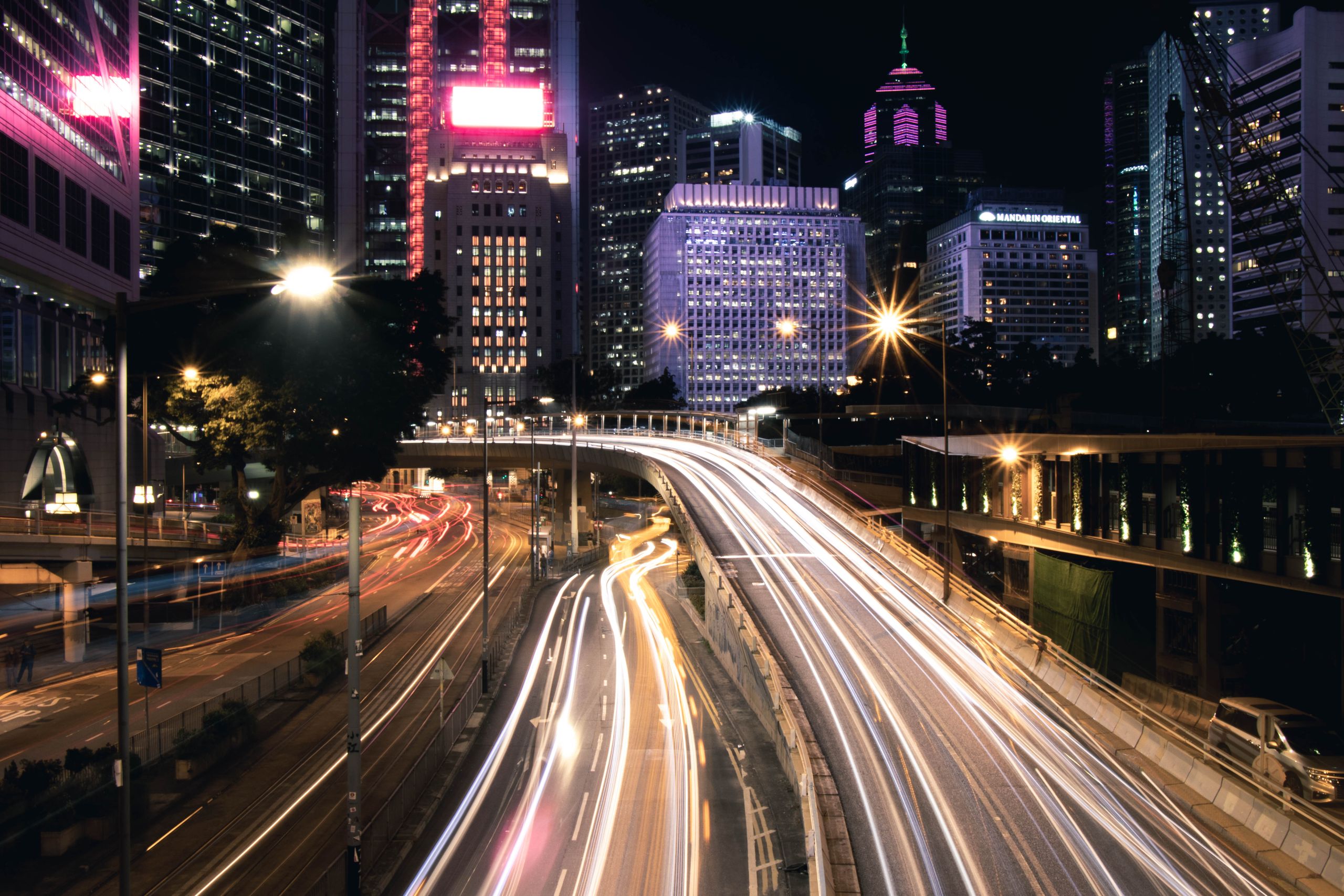 A city skyline at night with lit towers above a curving road streaked with light trails from traffic.