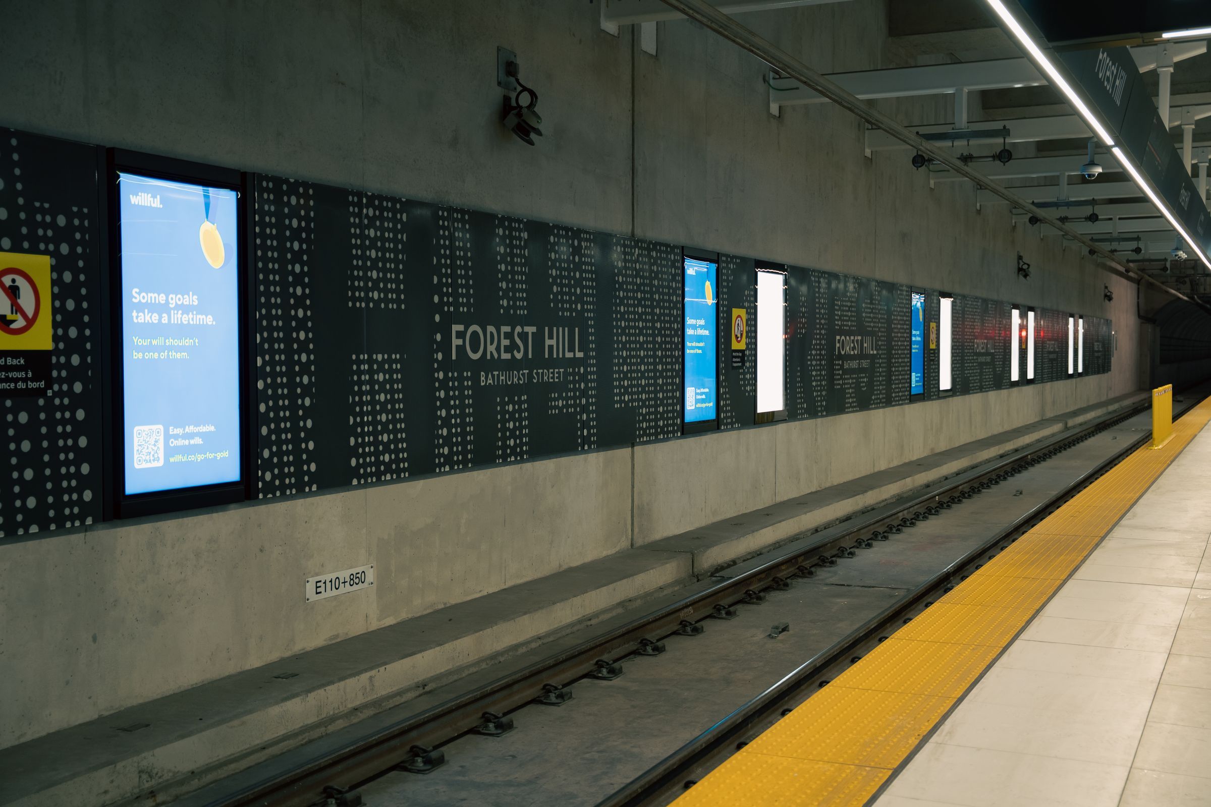 An underground metro platform with a yellow safety edge and a row of digital advertising and information screens along the wall.