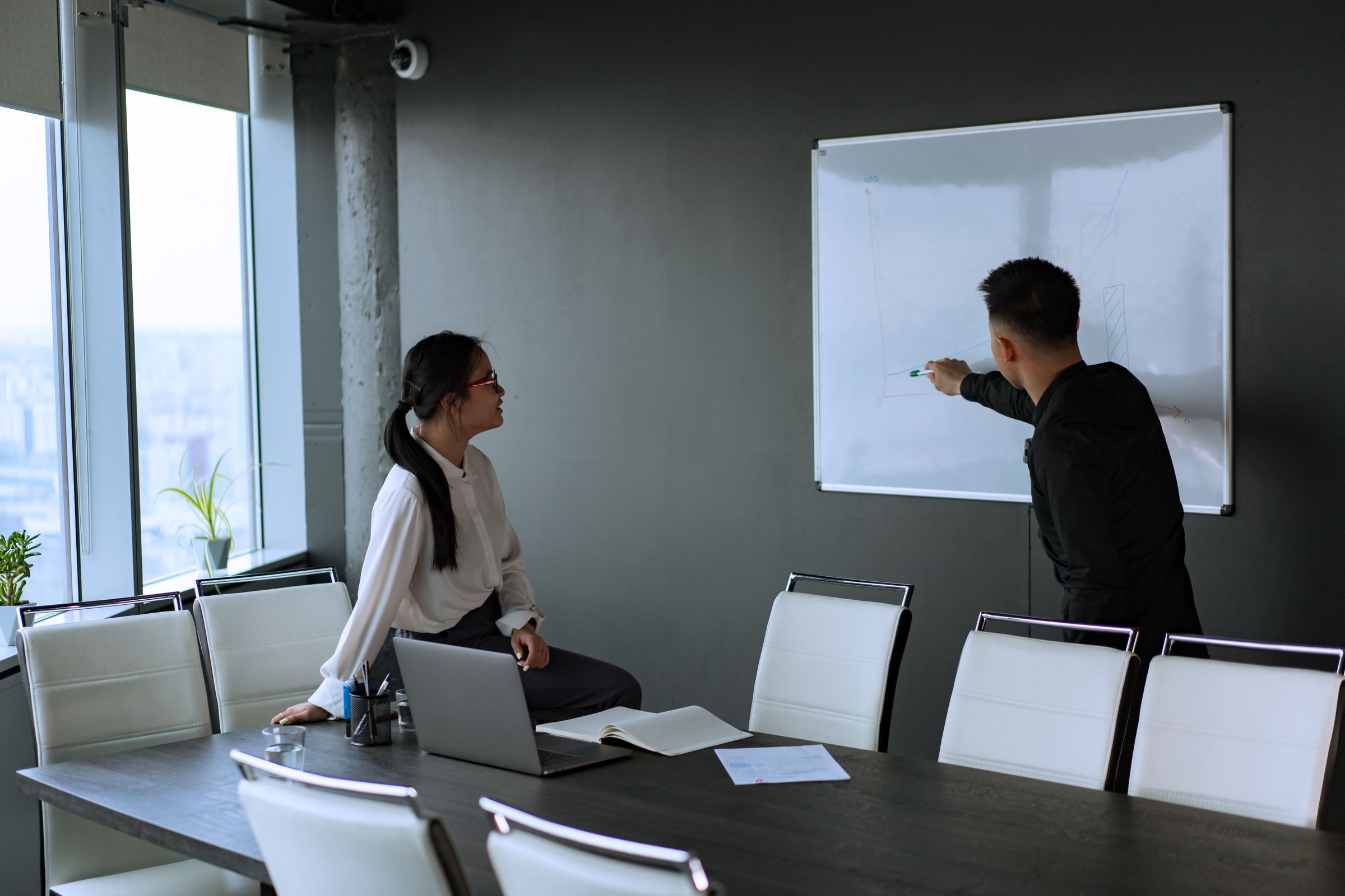 Two colleagues in a meeting room, one seated and one pointing at a whiteboard on the wall beside a long table.