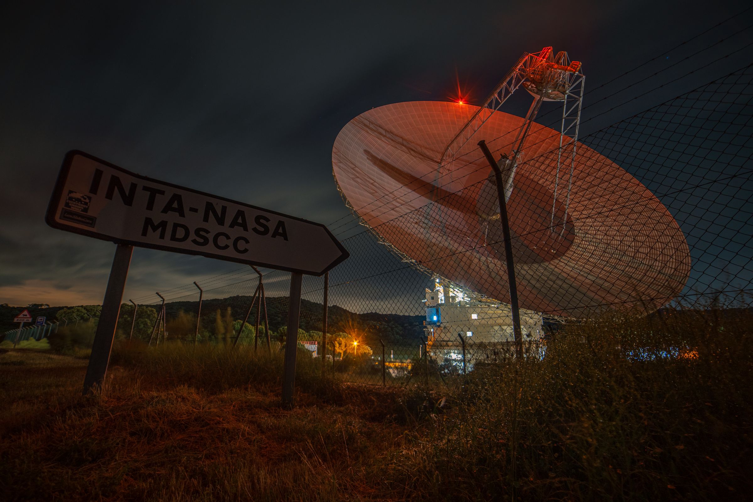 A large radio dish at night with a roadside sign in the foreground and a red light on its feed arm.
