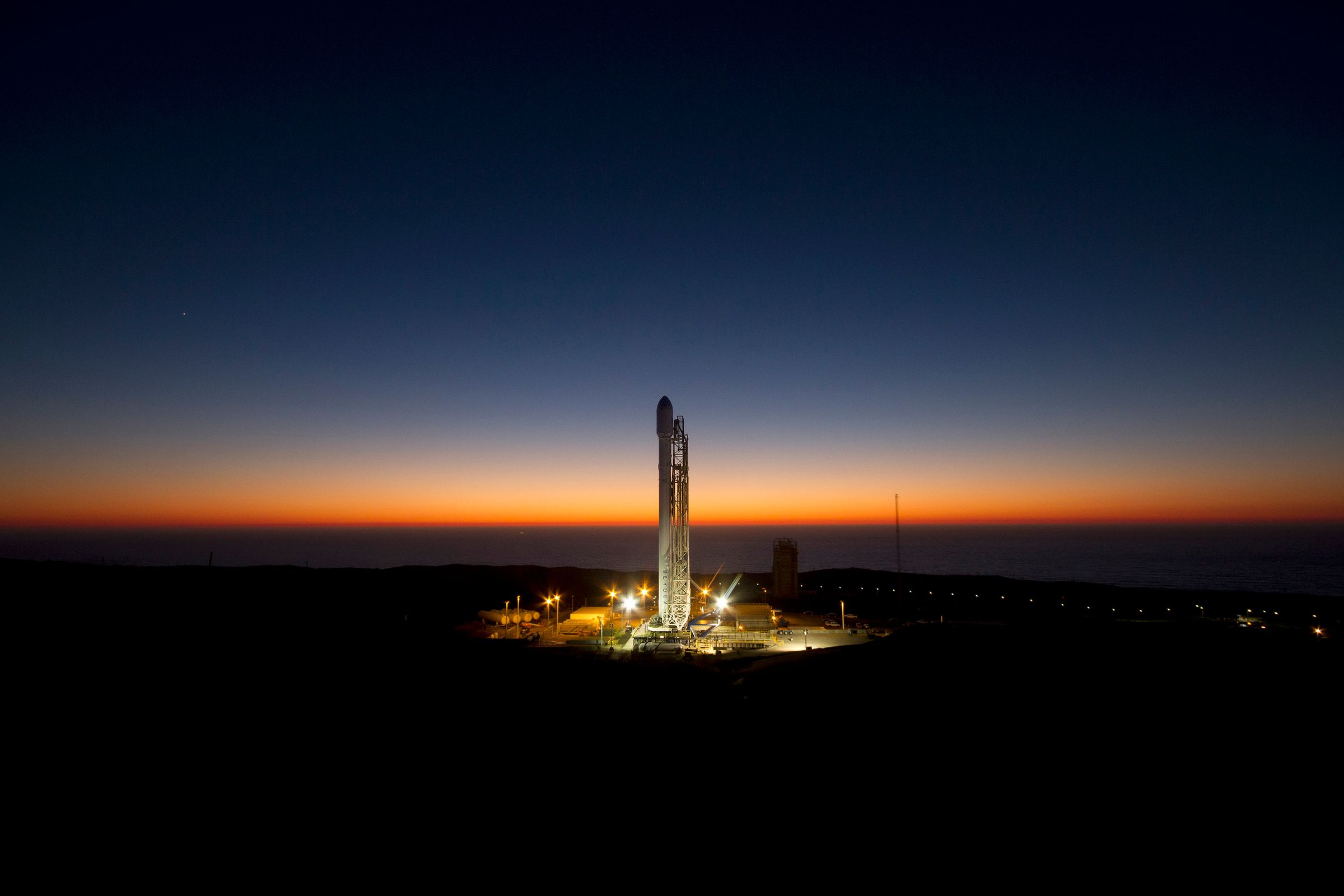 A rocket standing on a lit launch pad against an orange horizon at dawn.