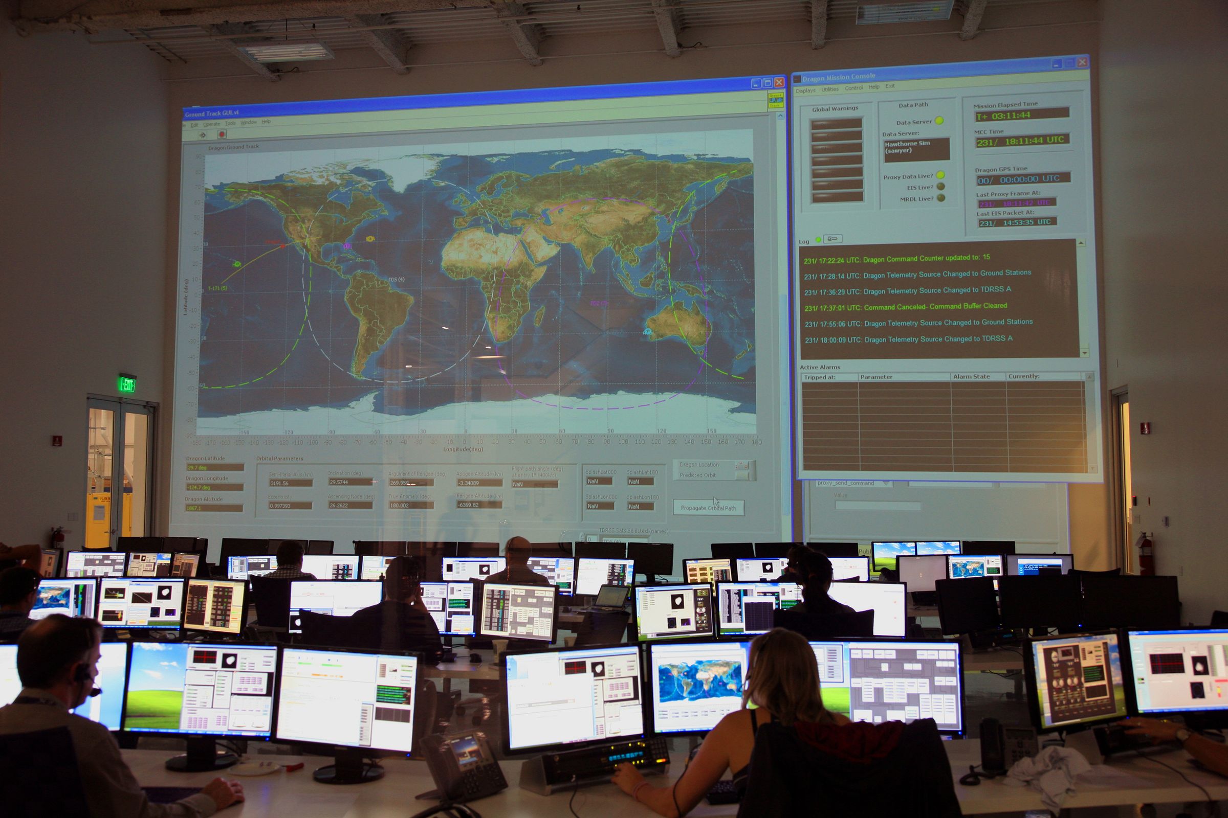 Operators at rows of monitors in a mission control room with a world map on a large screen.