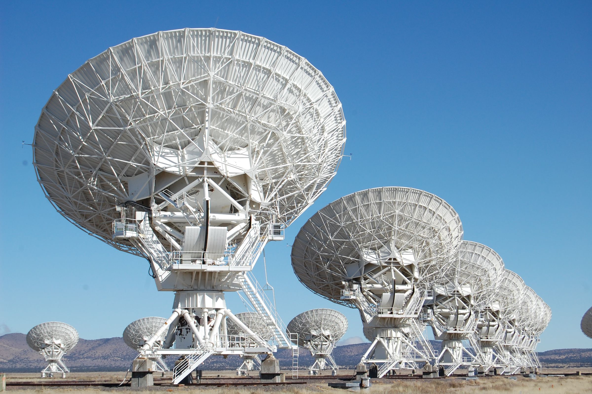 A row of large white radio telescope dishes in a dry landscape under a clear blue sky.