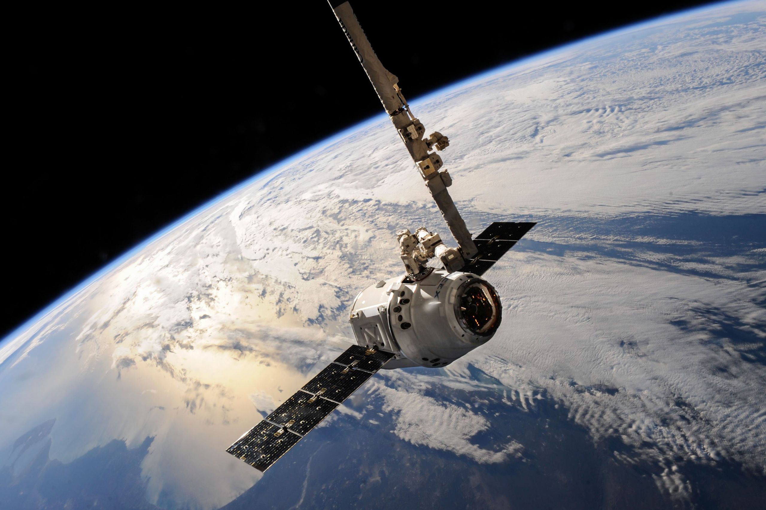 A spacecraft with solar panels and a robotic arm above the curved edge of Earth, with white cloud below.