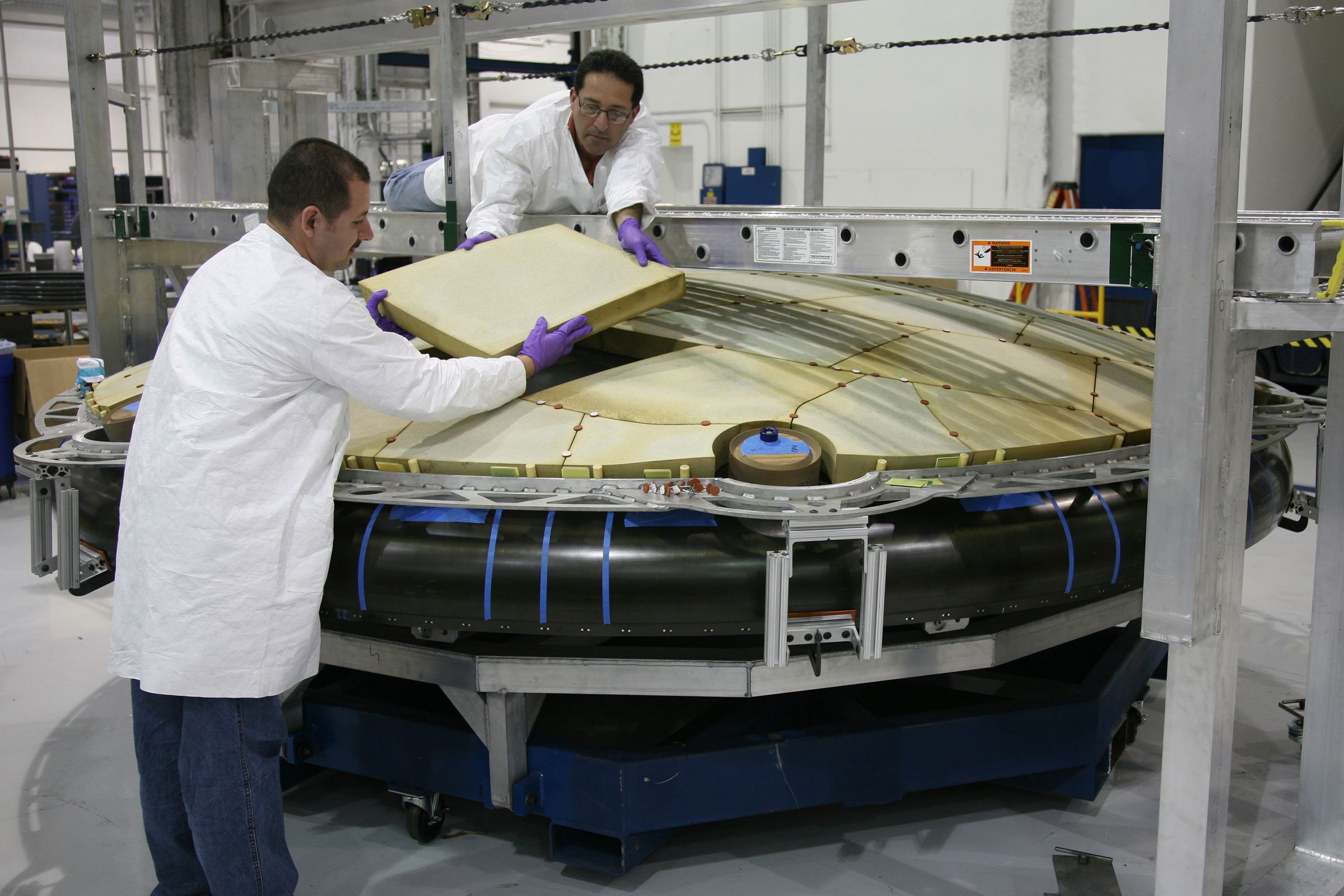 Two technicians in white coats lifting a panel on a large spacecraft structure in a hangar.