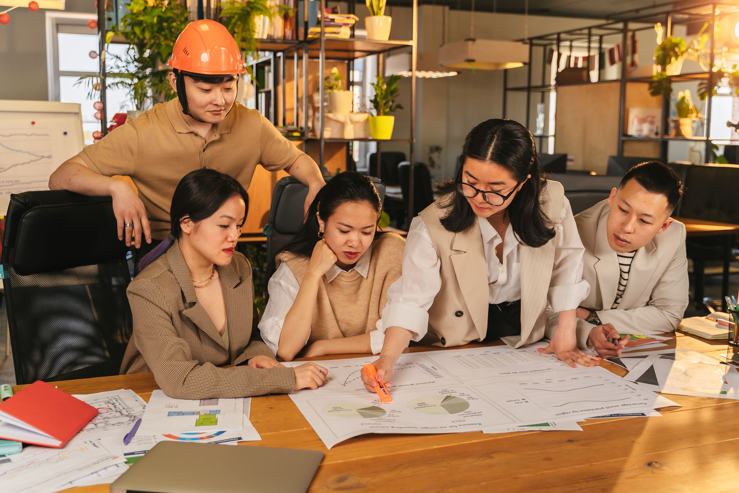 Five colleagues, one in an orange hard hat, leaning over plans spread across a wooden table.