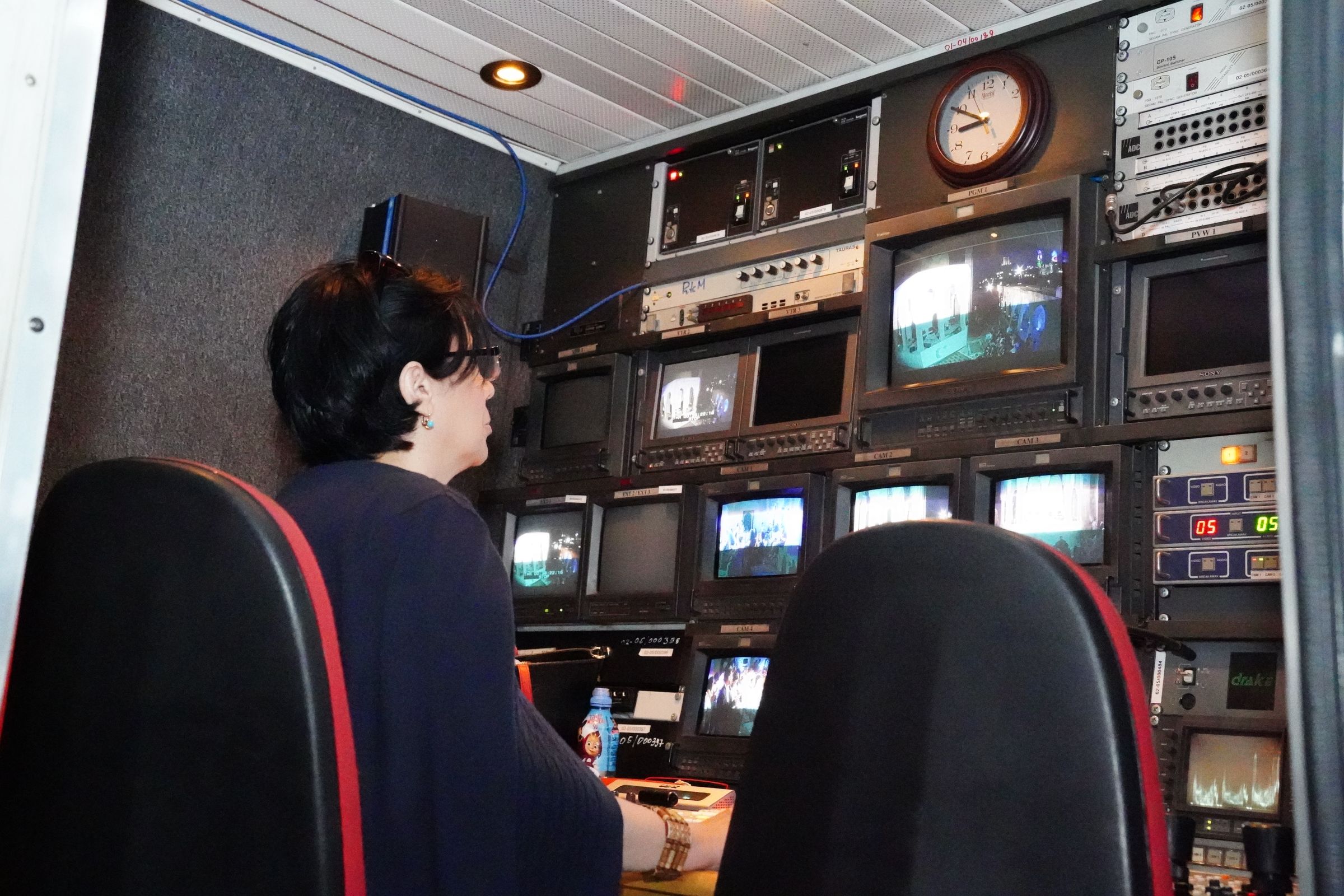 A technician seated at a broadcast control desk in front of racks of video monitors and switching equipment.