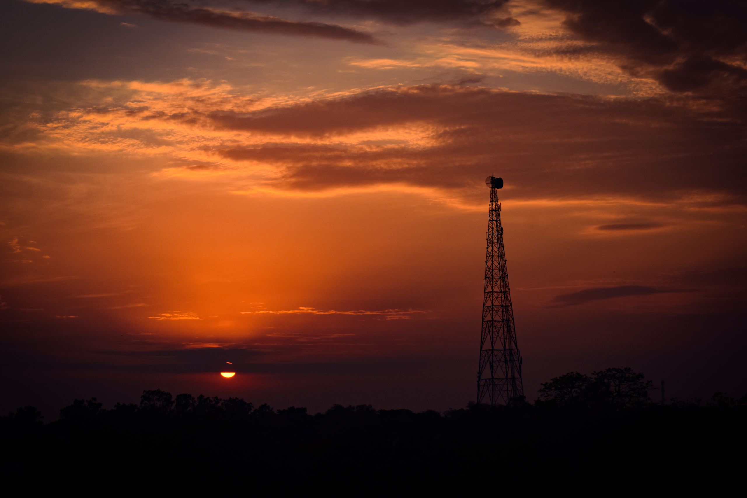 A lattice telecoms mast silhouetted against an orange sunset sky with low cloud.