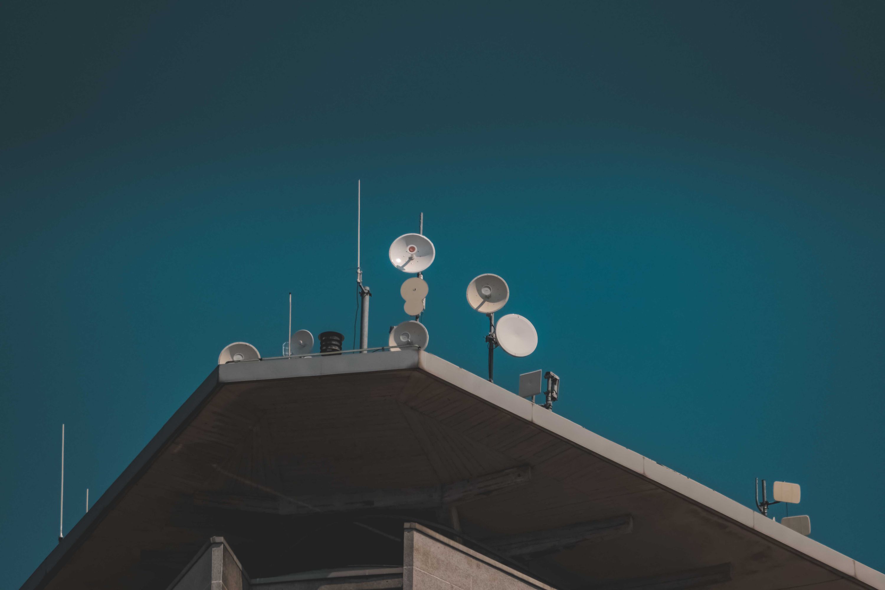 Microwave dishes and antenna poles mounted on the ridge of a rooftop against a deep teal sky.