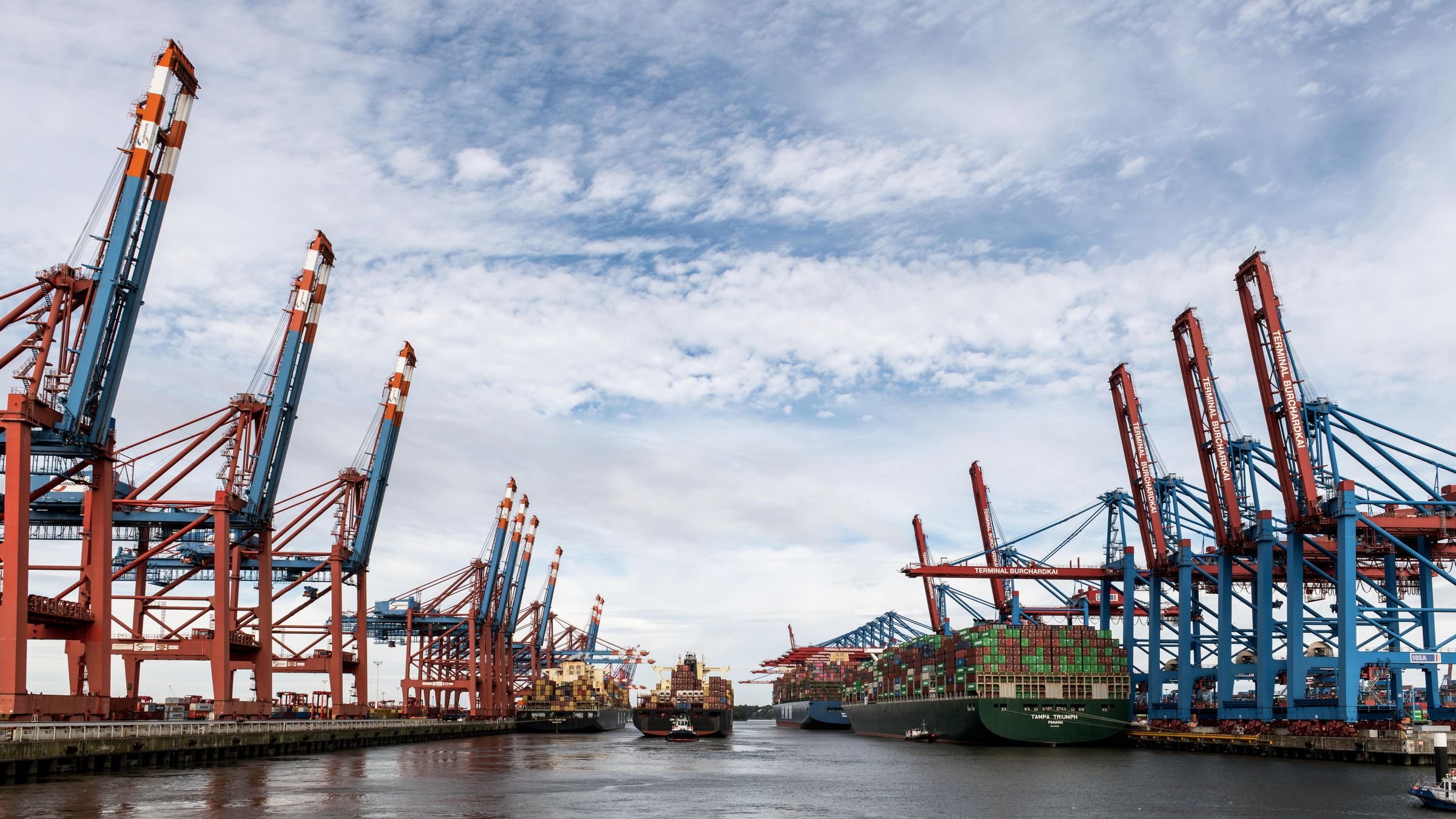 Container cranes and ships at a busy port under a cloudy sky
