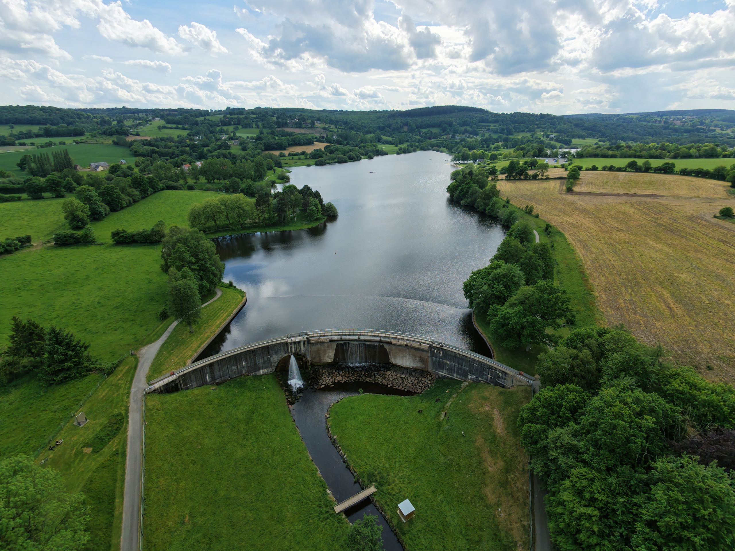 An aerial view of a dam and narrow reservoir among green fields and woodland under a cloudy sky.