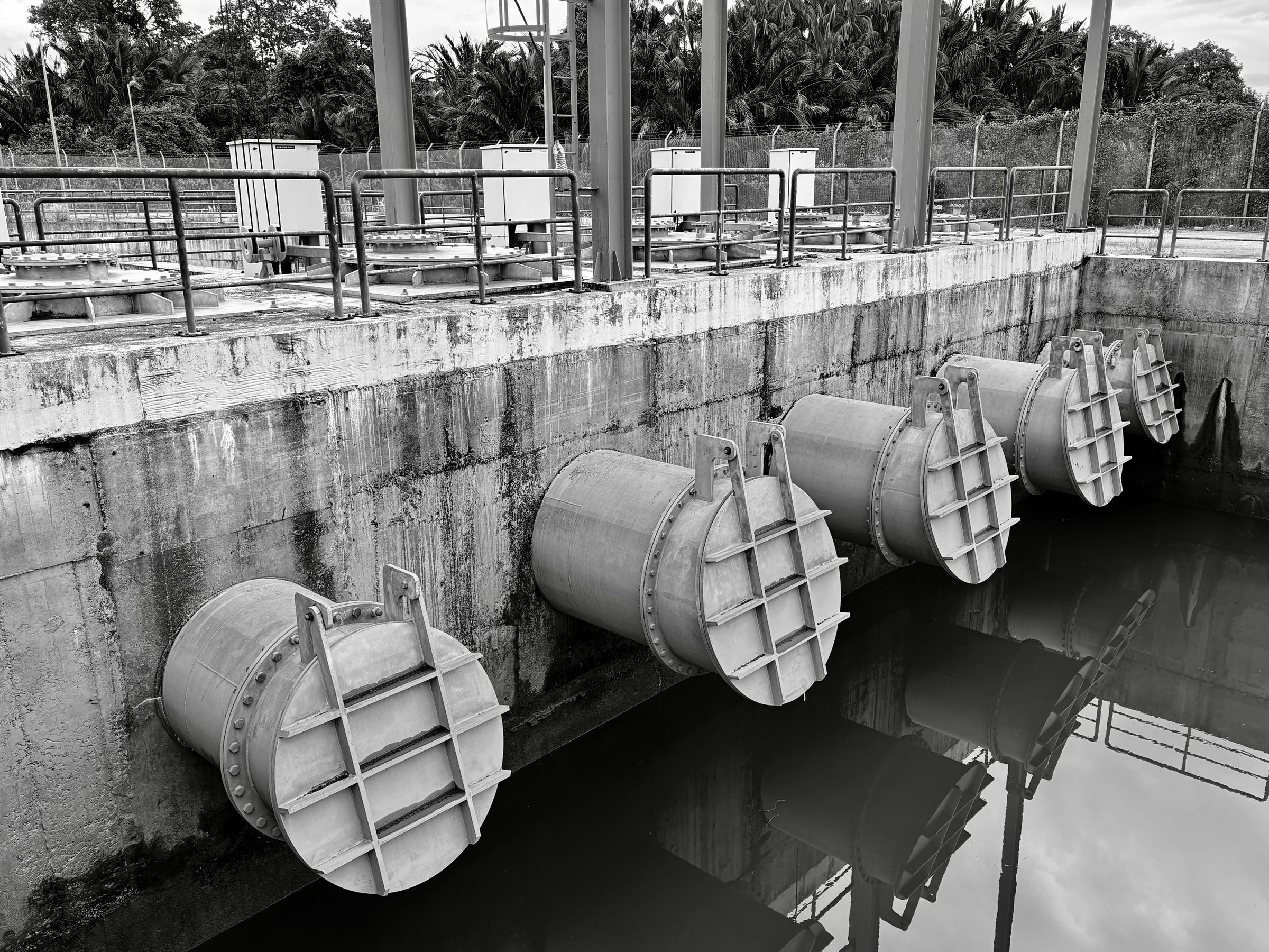 Large steel pipe outlets and sluice gates set into a concrete wall above dark water, in black and white.