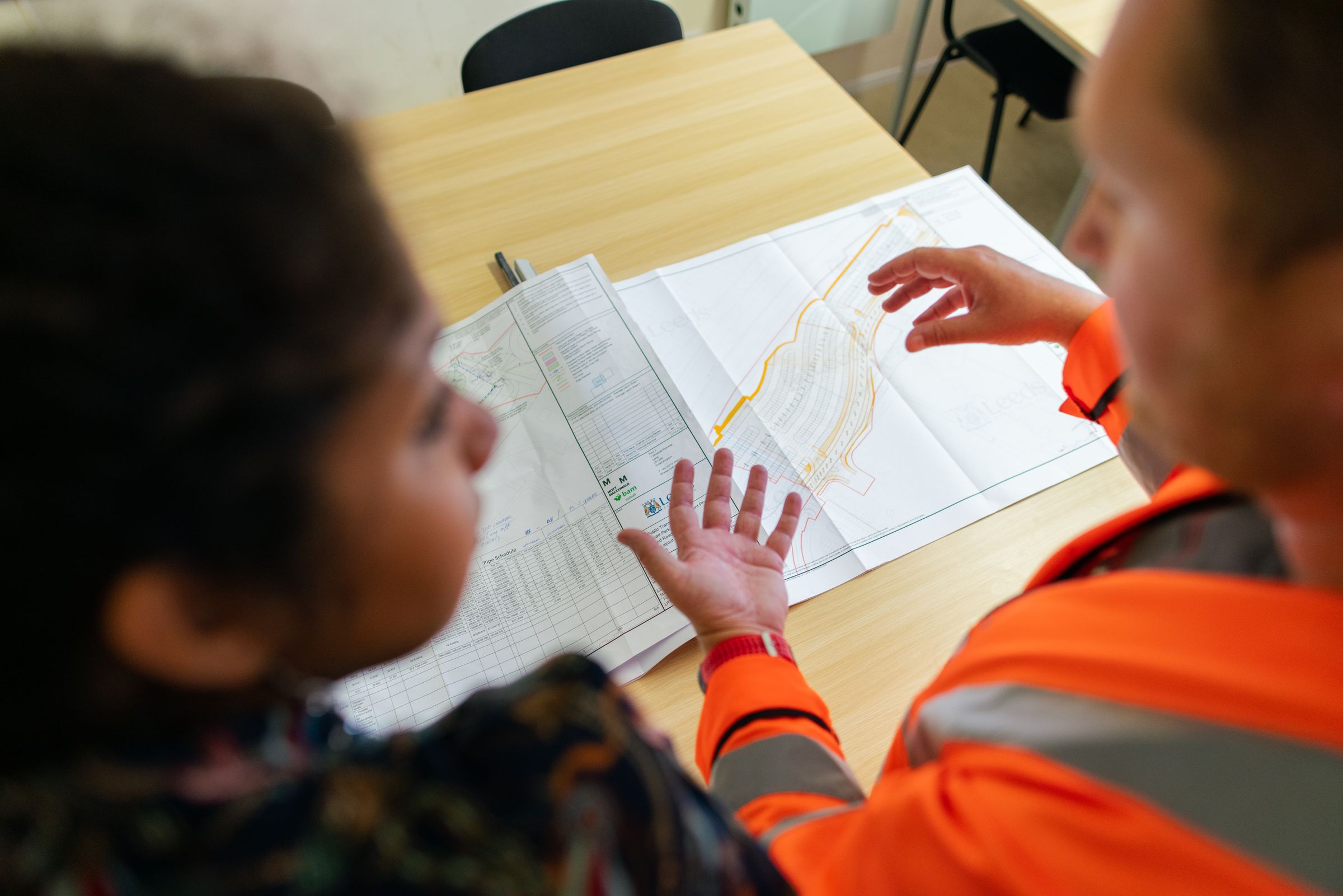 Two people in high-visibility clothing pointing at large site plans spread on a table, seen from above.