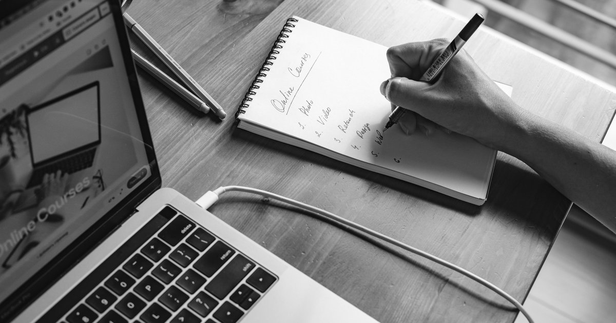A hand writing a numbered list in a notebook beside a laptop open to an online courses webpage