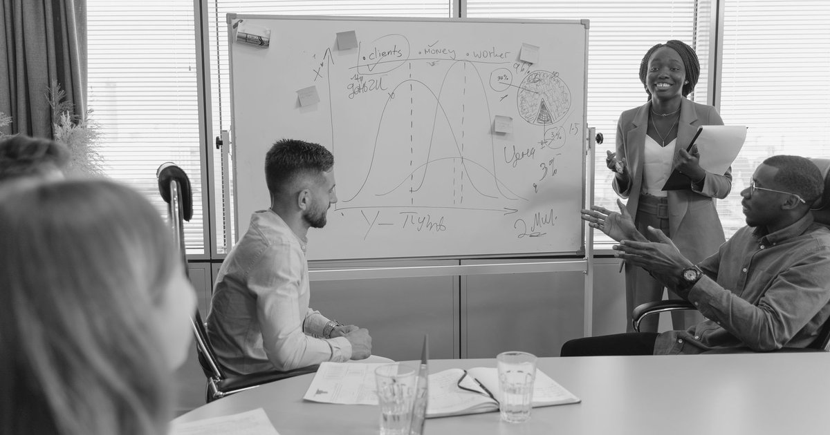 A presenter at a whiteboard explaining a chart to three colleagues in a meeting room