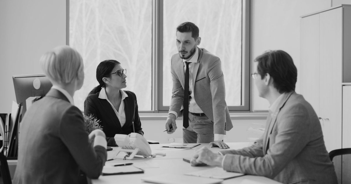 Four executives in discussion around a boardroom table, one standing and explaining a point